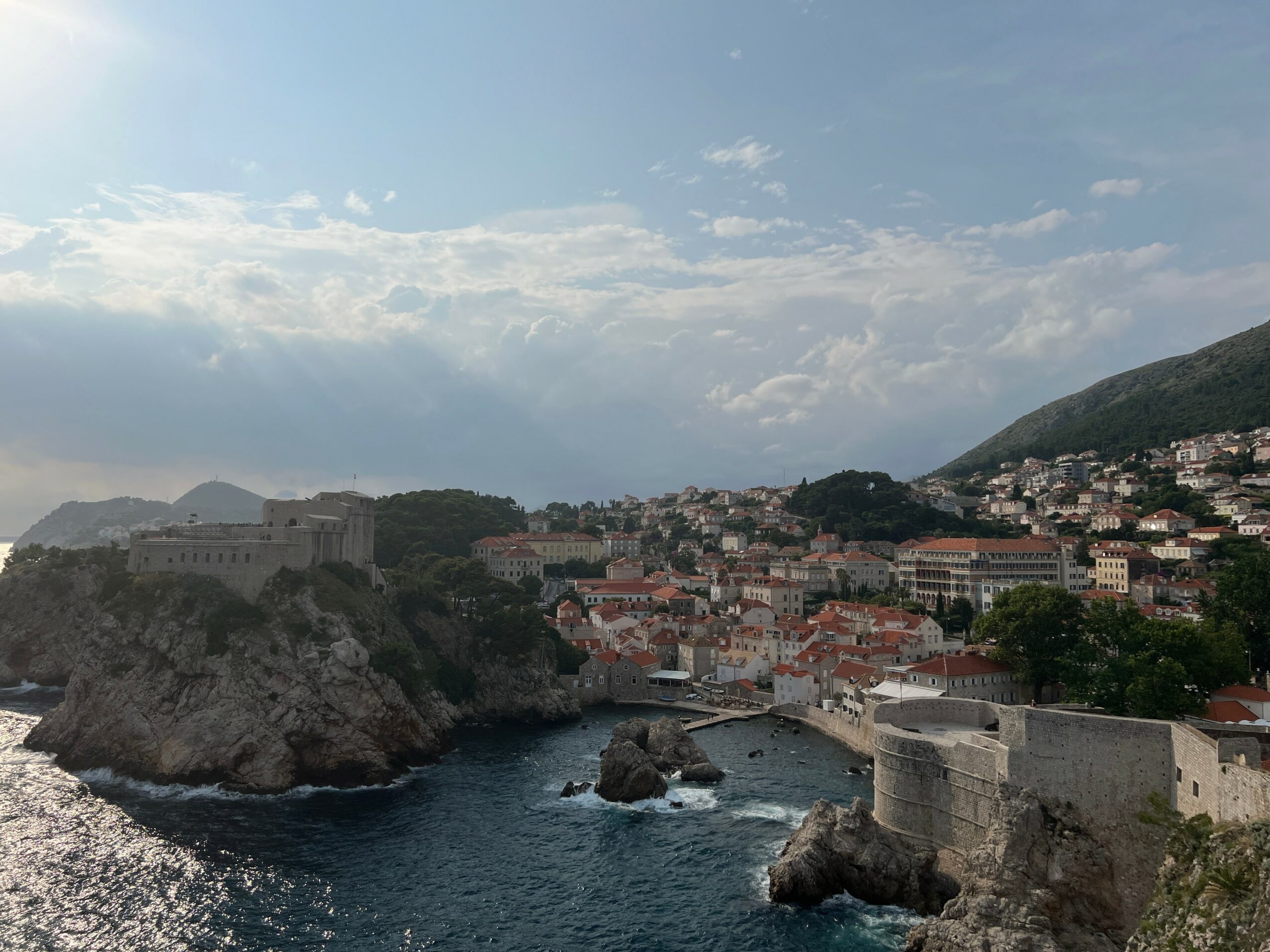 Coastal city with buildings on hills, a rocky shoreline, and a body of water under a partly cloudy sky.