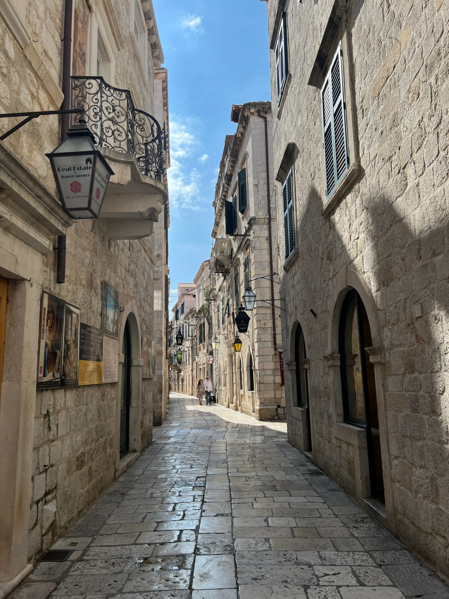 Narrow cobblestone street lined with stone buildings, some with balconies and windows, under a partly cloudy sky.