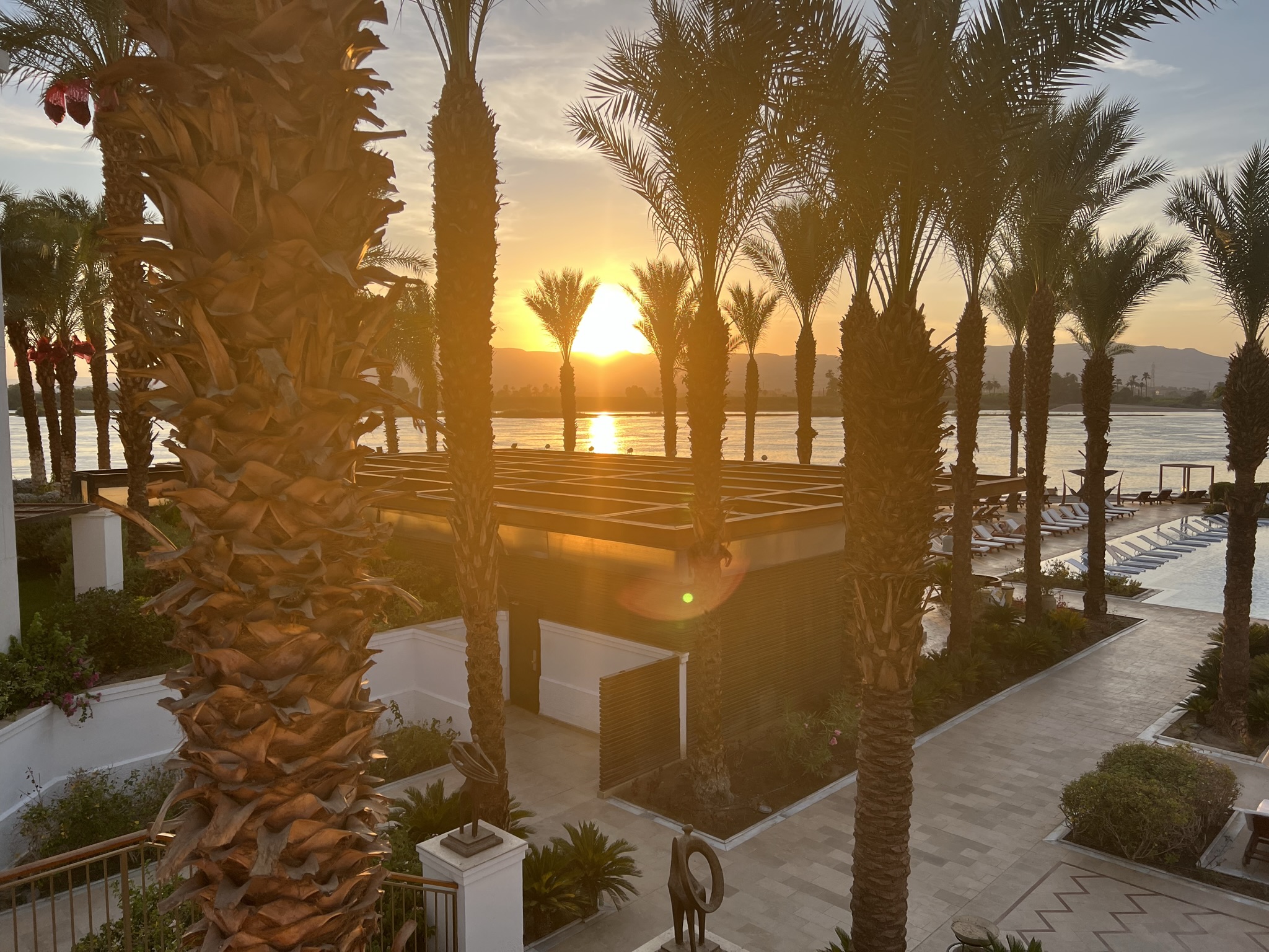 Sunset over a waterfront with palm trees and a walkway, with a small building in the foreground.