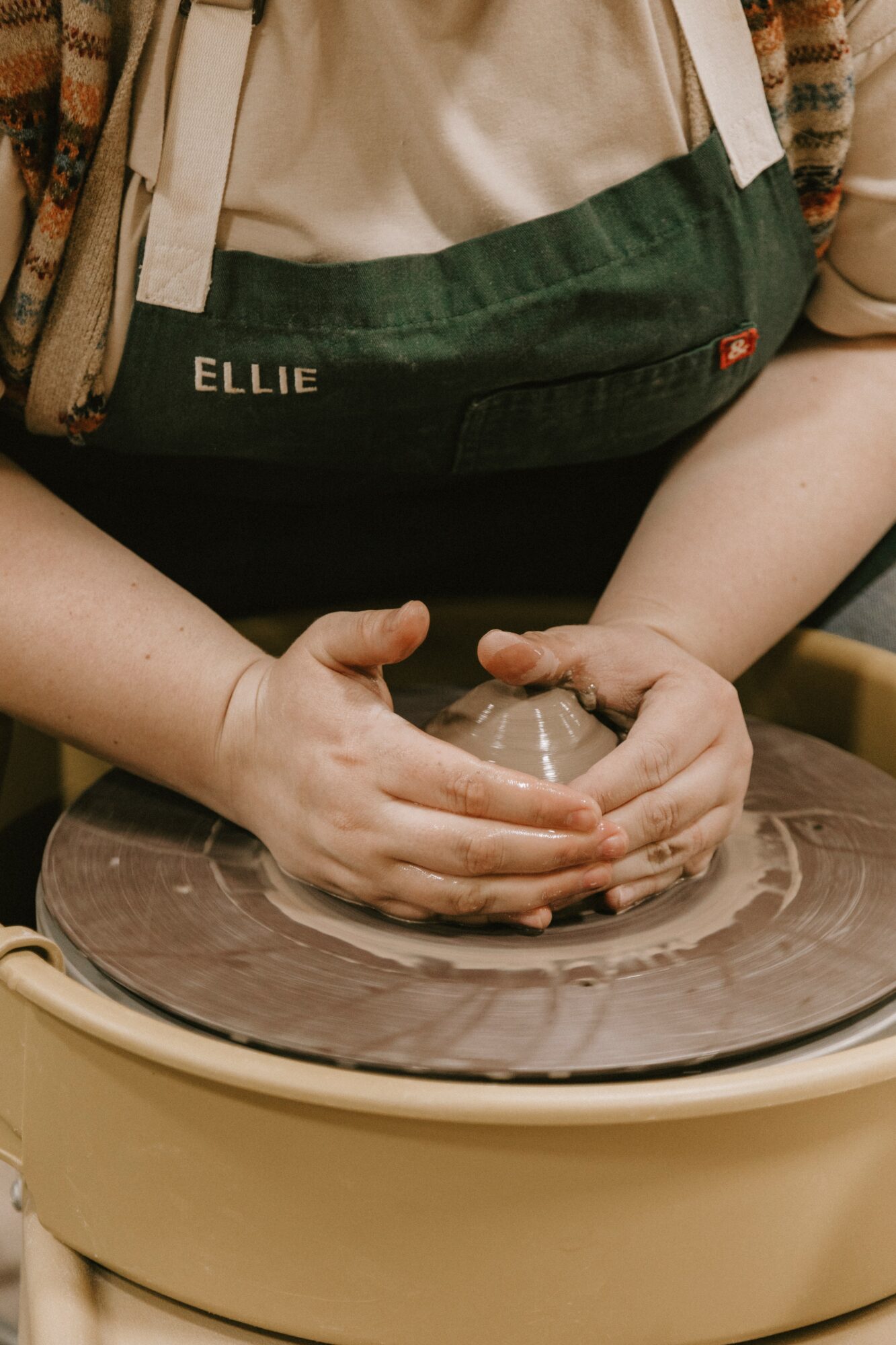 Person shaping clay on pottery wheel with hands, wearing an apron with the name 'Ellie'.