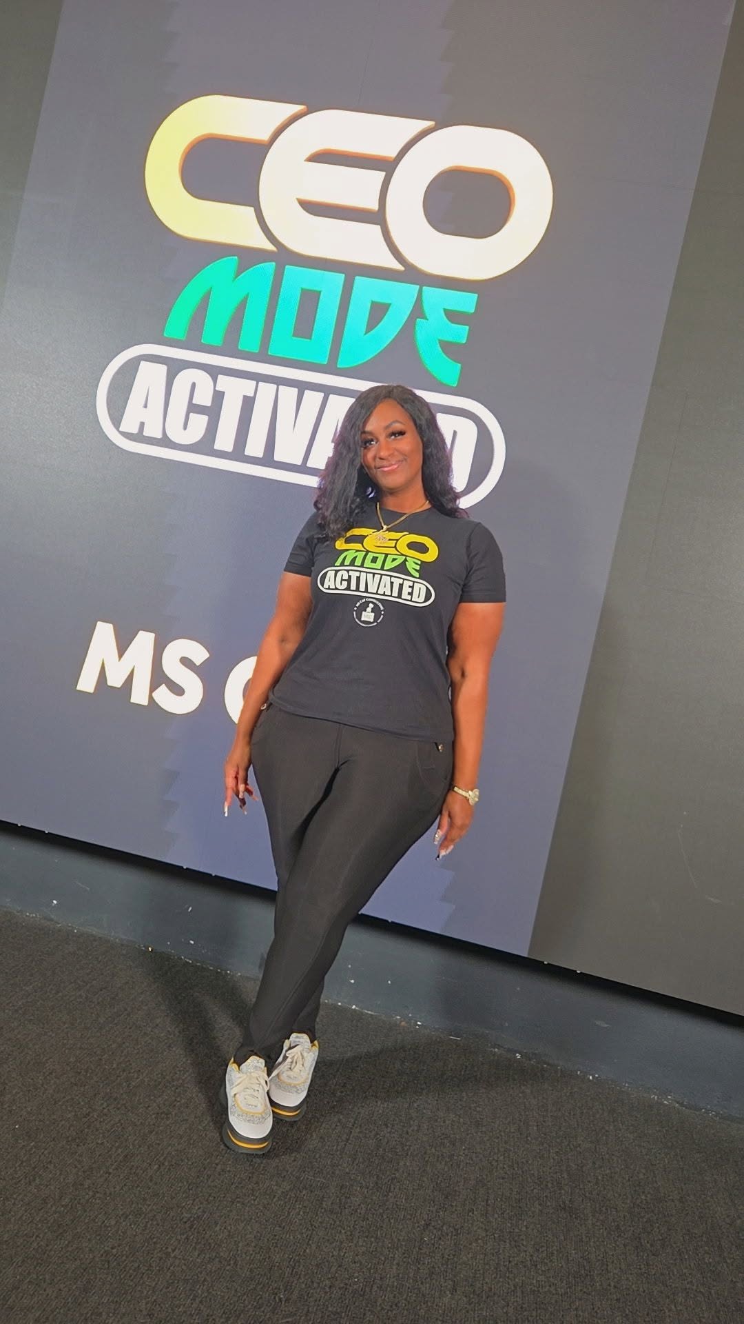 Woman standing in front of a large screen with text and logos, wearing a black T-shirt and gray pants.