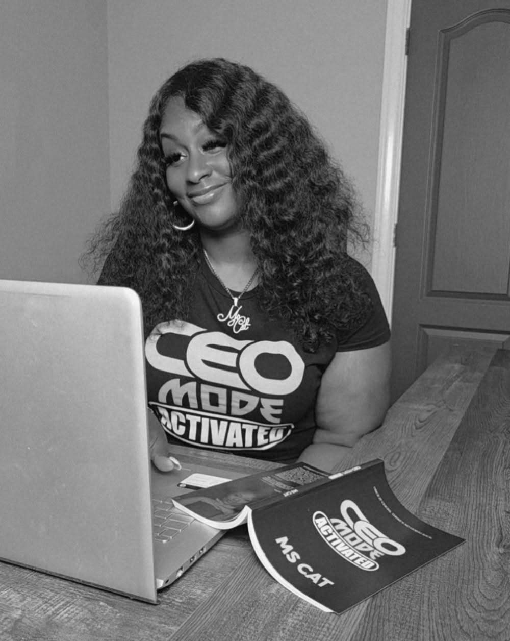 Woman with curly hair sitting at a table with a laptop and papers, smiling, in black and white.
