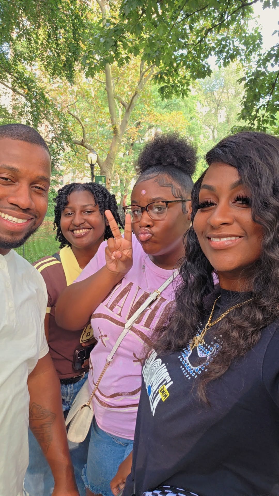 Four people smiling outdoors with trees in background, one making a peace sign, all wearing casual clothing.