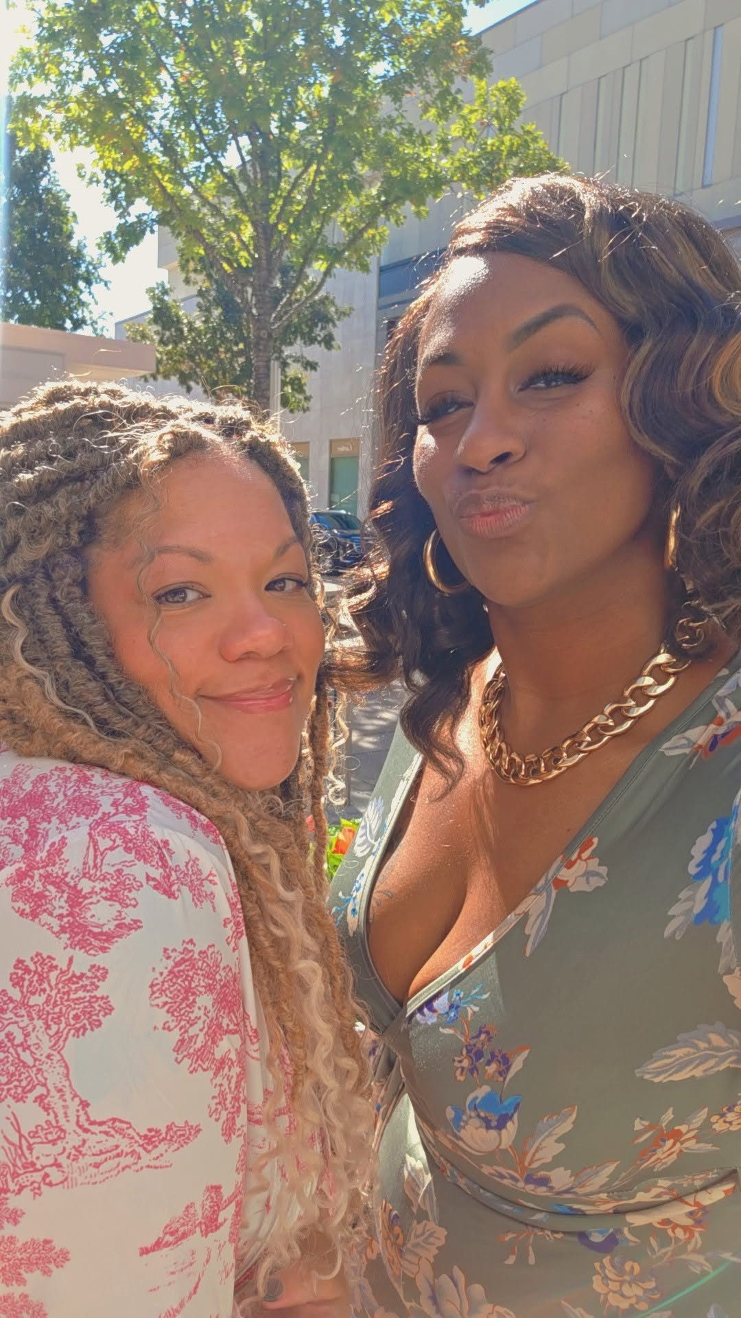 Two women taking a selfie outdoors, smiling and making playful expressions, with trees and a building in the background.