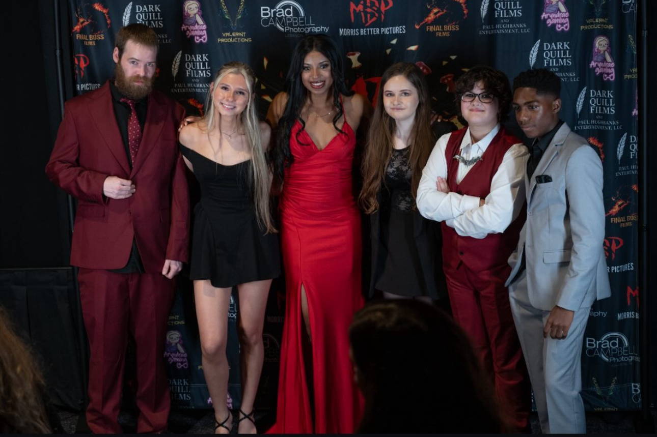 Group of six people standing together on a stage with a dark backdrop, some smiling and dressed formally.