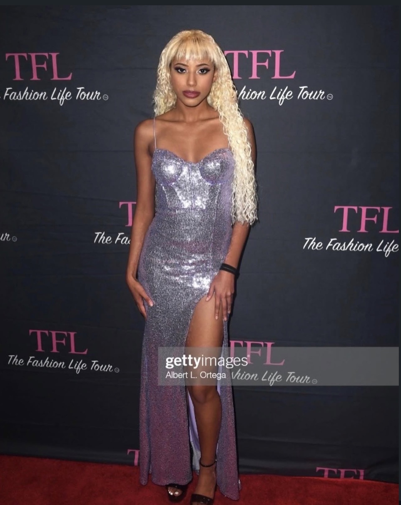 Woman in a silver sequined dress with a high slit, standing on a red carpet at an event, with a dark background.