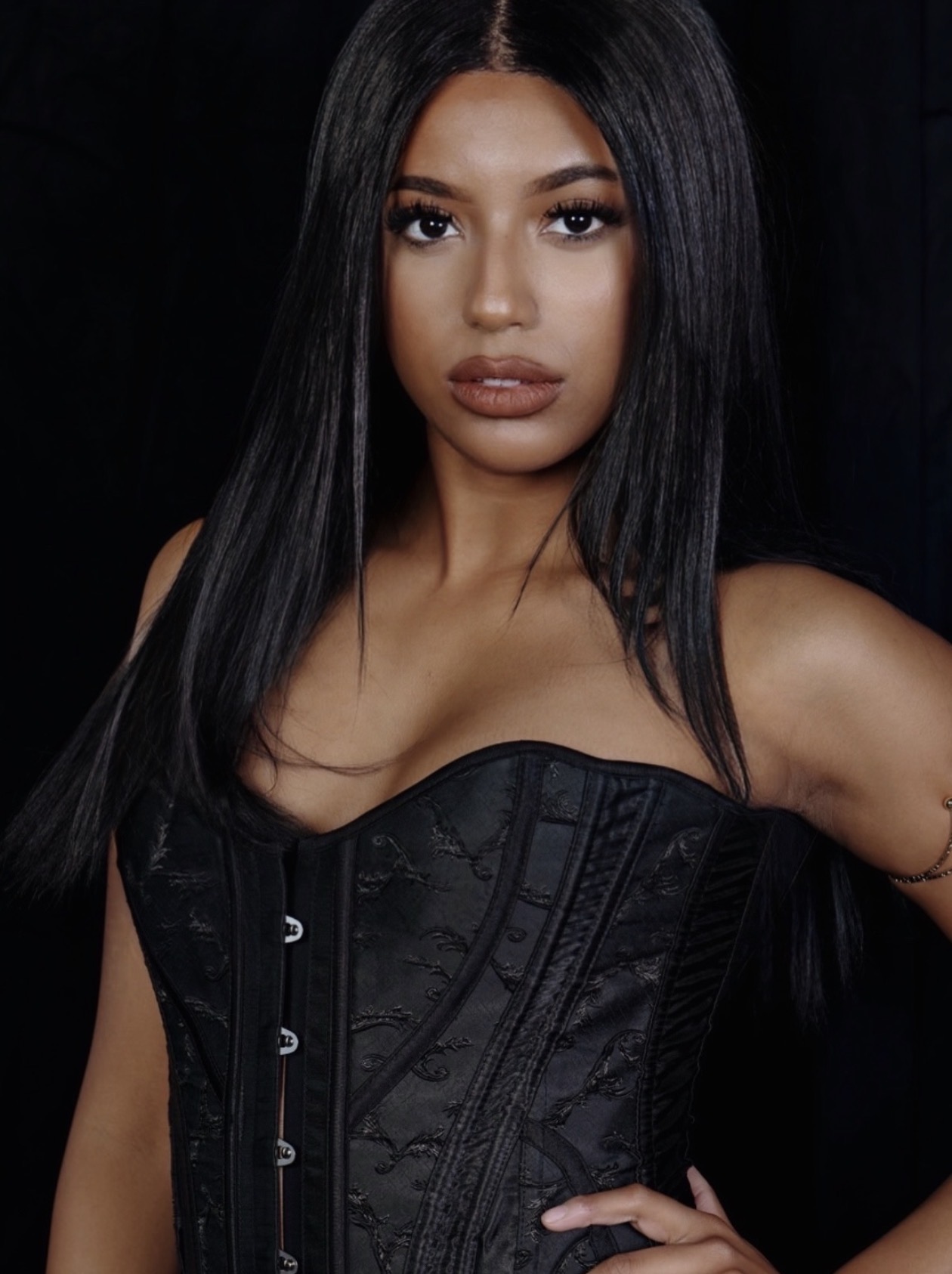 Young woman with long black hair wearing a black corset top, posing against a dark background.