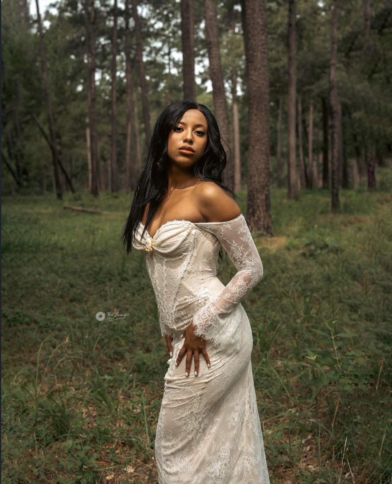Woman in a white off-shoulder dress standing in a forest with trees and greenery.