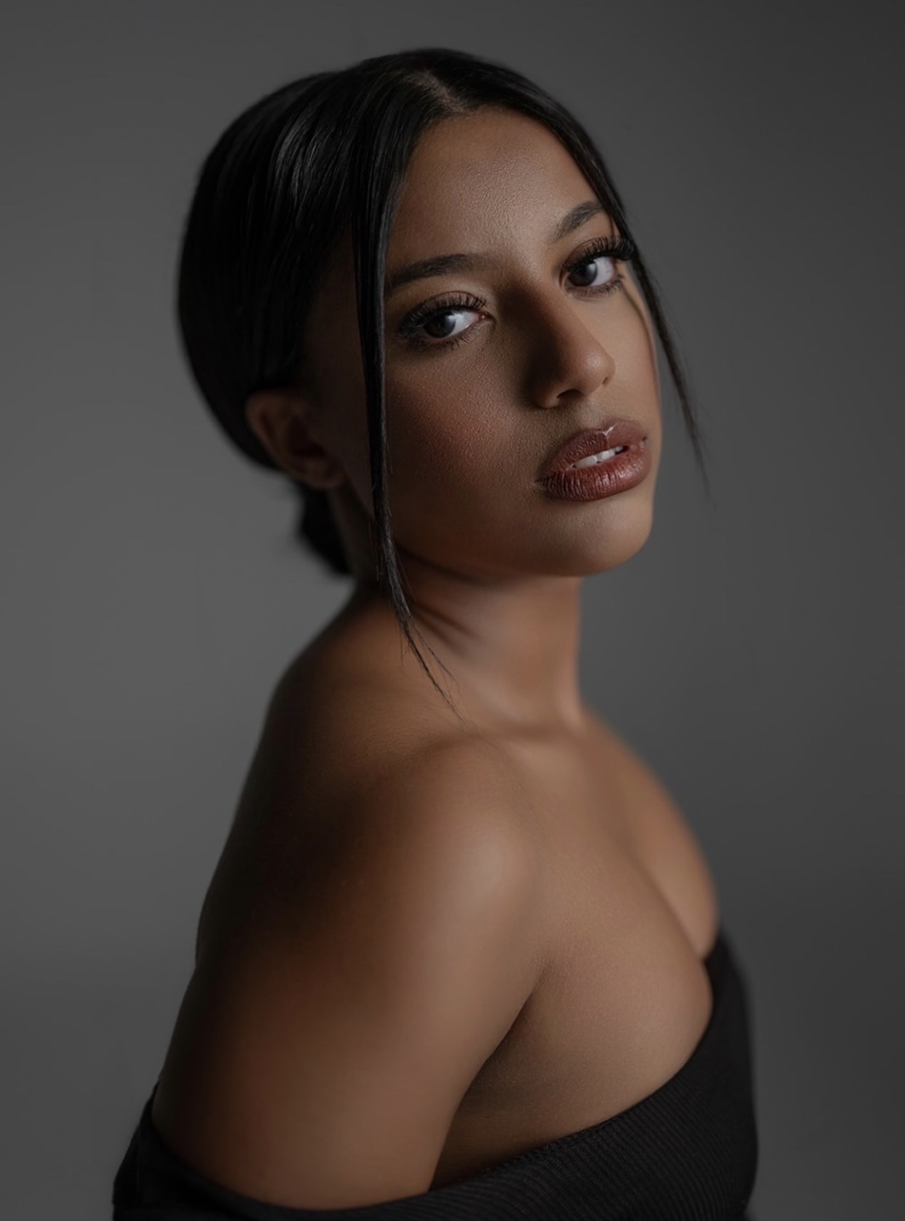 Portrait of a woman with short black hair, wearing a strapless black top, looking at the camera against a dark background.