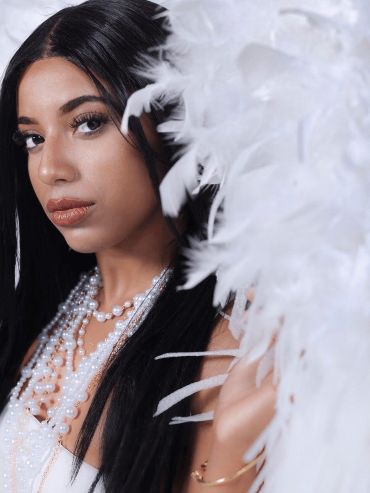 Close-up of a woman with long dark hair, wearing pearl necklaces and a black outfit, holding white feathers.
