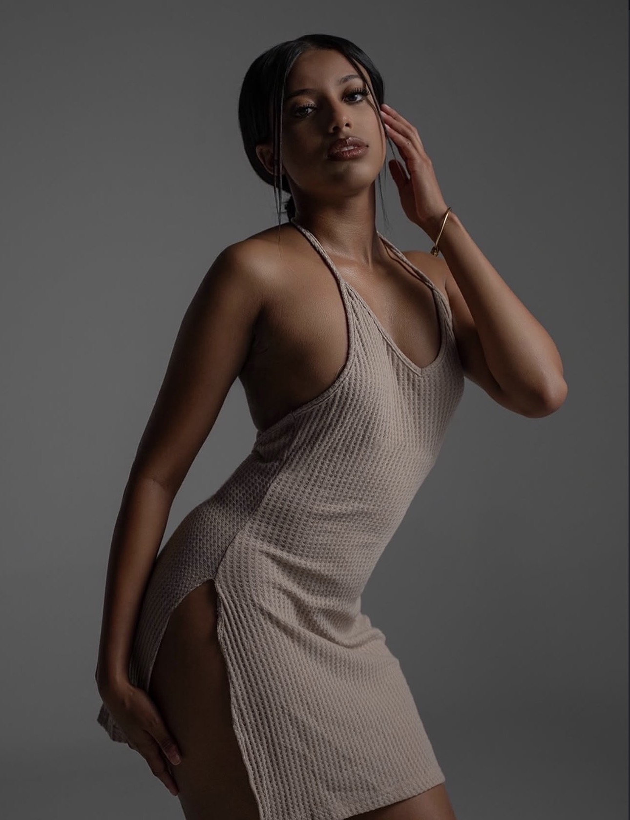 Woman in beige dress poses with hand near face against gray background.
