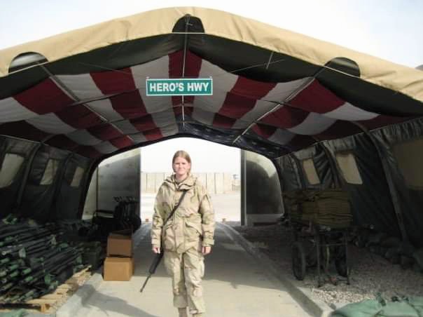 Person standing under a tent with a street sign reading 'Hero's Hwy' above. The person is holding a tool and wearing a jacket.