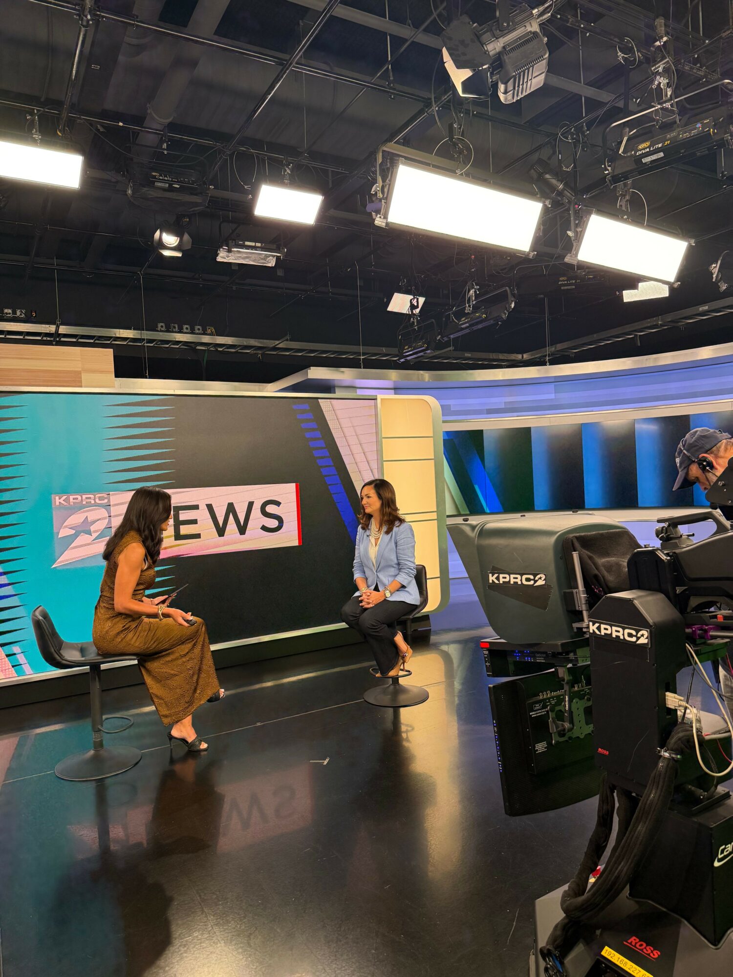 Two women sit on chairs in a television studio with a large screen displaying the name 'REBEWS' behind them.