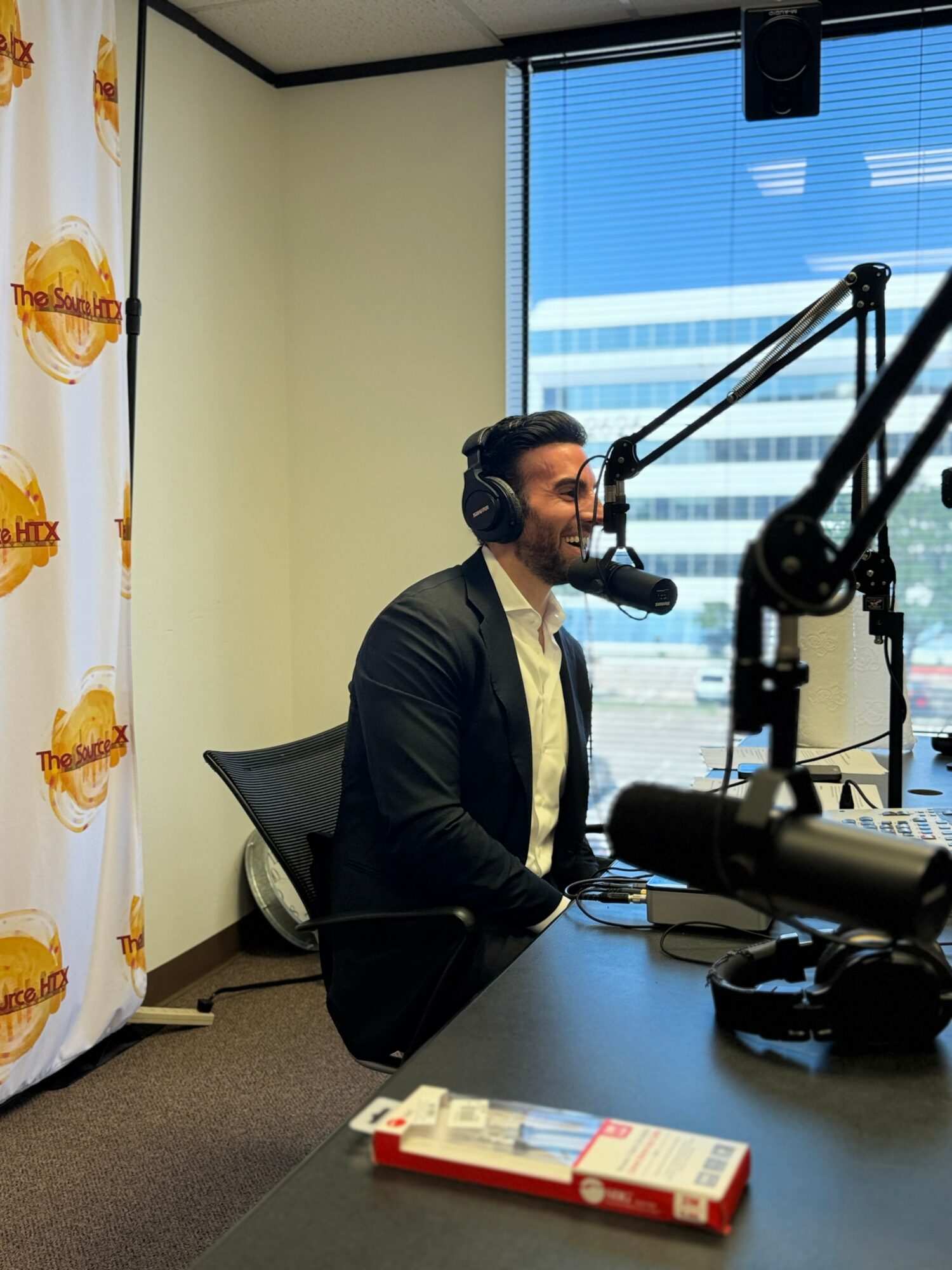 Man wearing headphones speaking into a microphone in a radio studio with a window showing a cityscape.