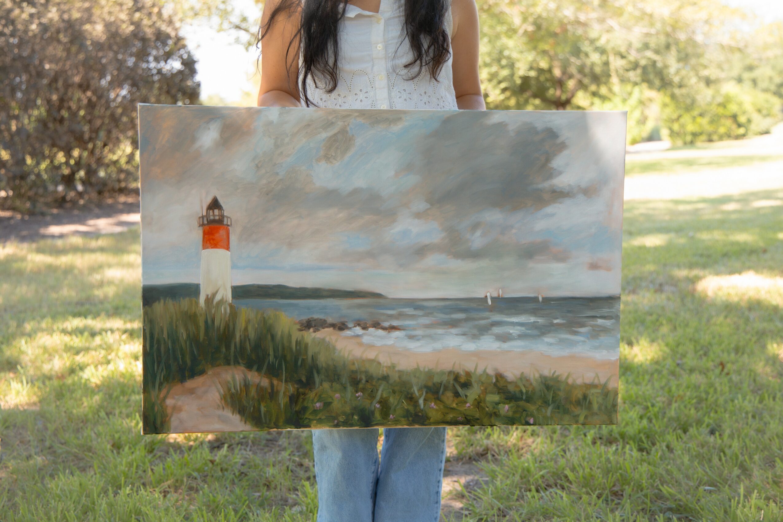 Person holding a landscape painting of a lighthouse, water, and sky outdoors, with trees and grass in the background.
