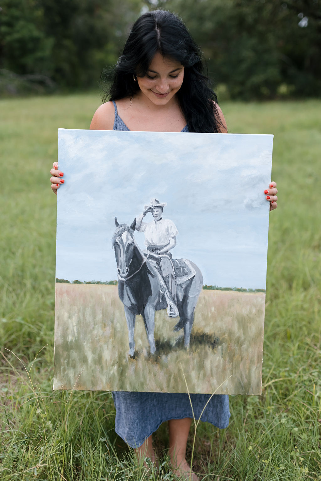 Girl holding a painting of a person riding a horse outdoors in a grassy field with trees in the background.