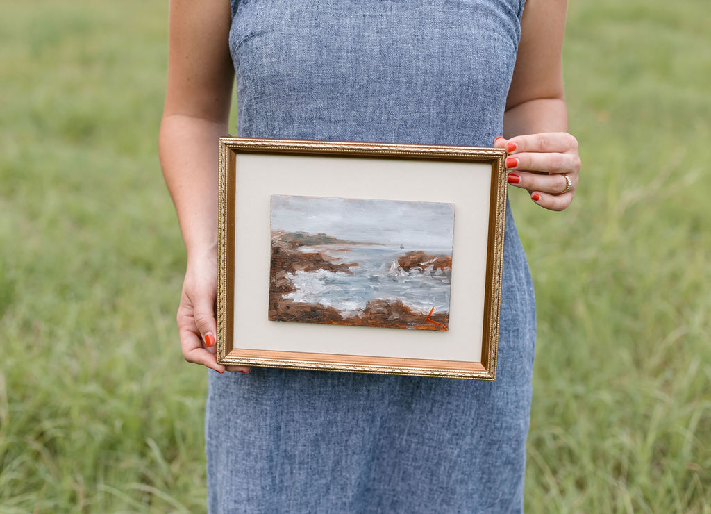 Person in a blue dress holding a framed landscape painting outdoors, grass background.