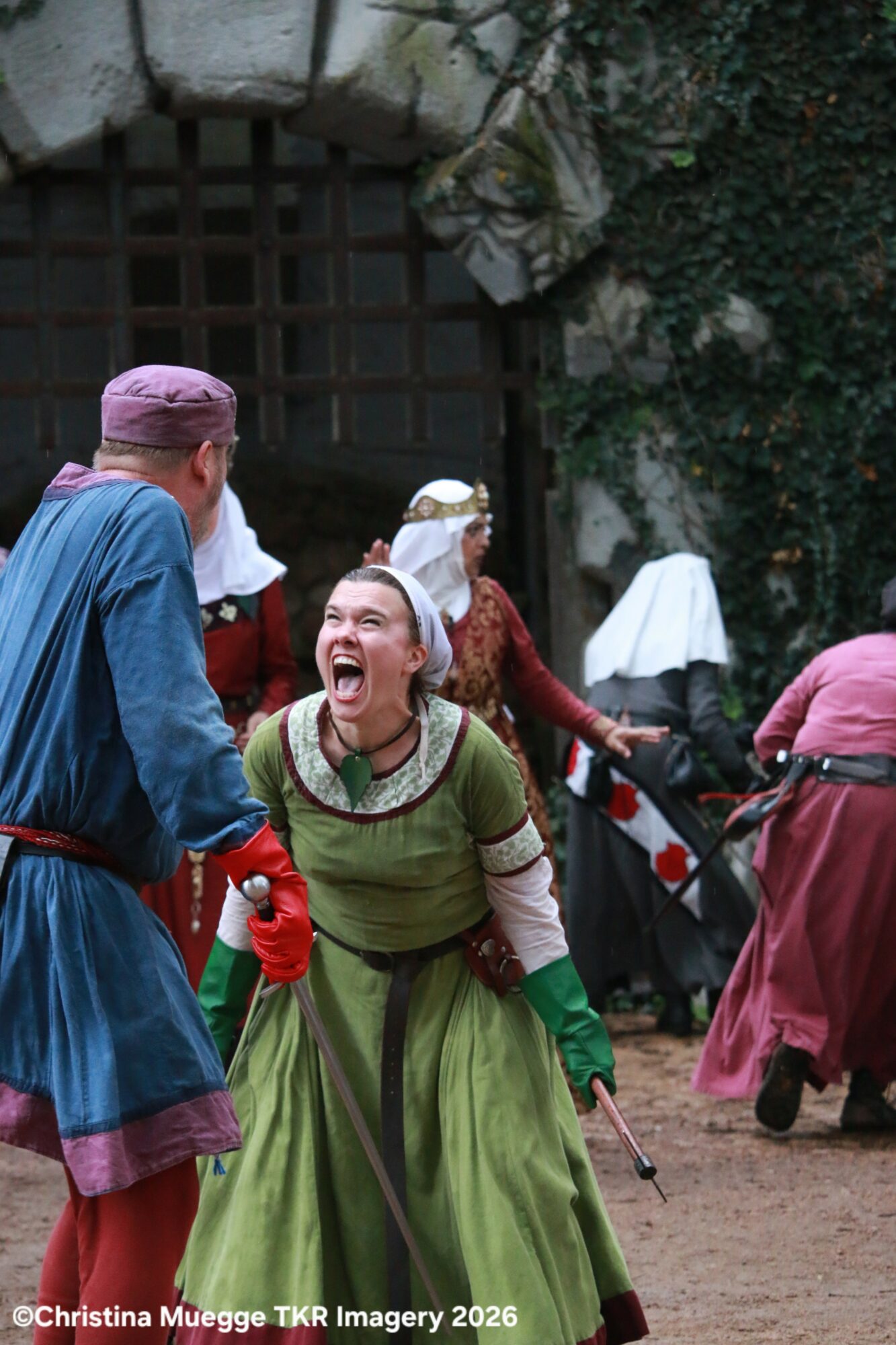 Two people in medieval costumes laughing and talking outdoors, with others in similar attire in the background.