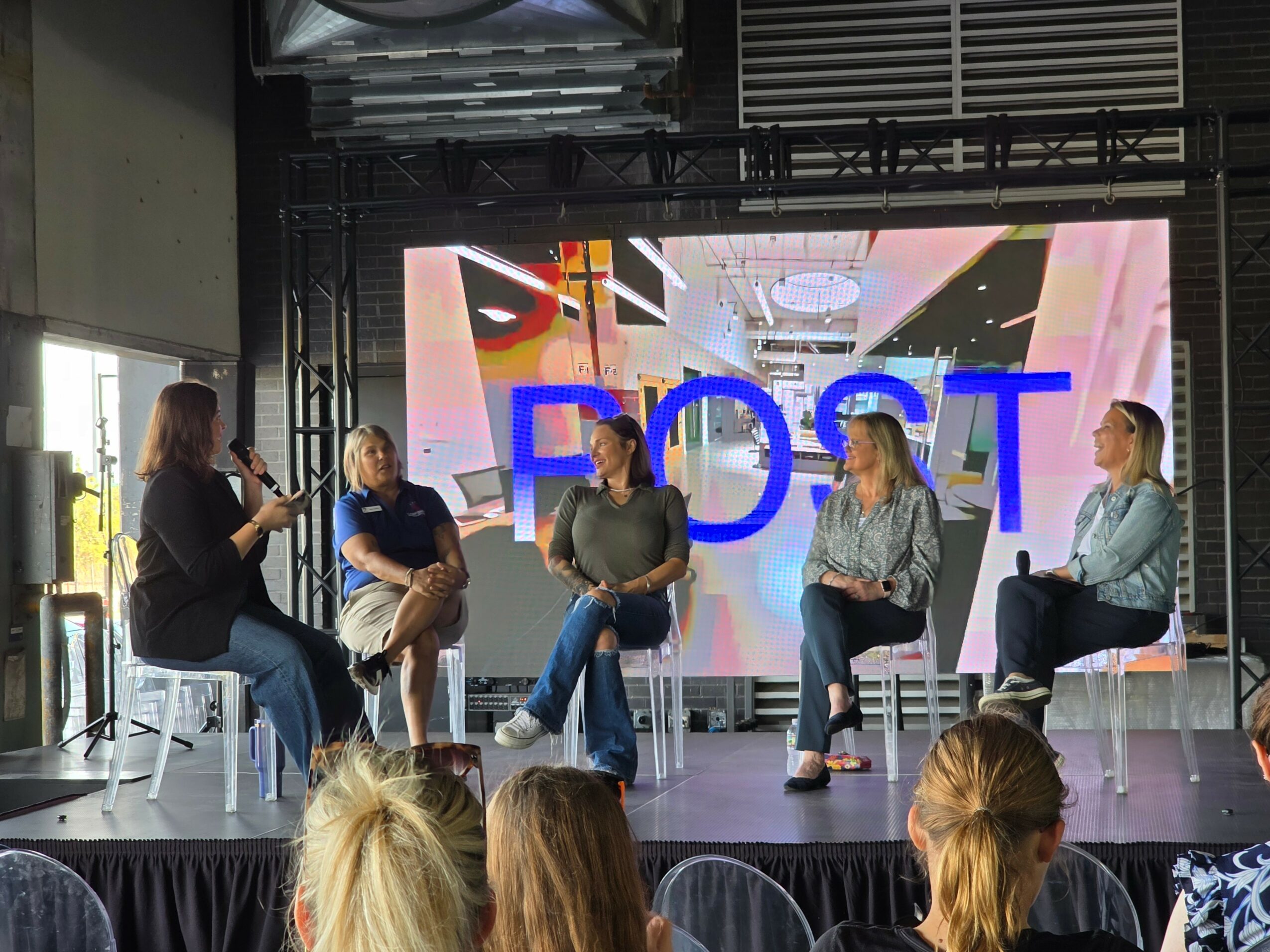 Five women seated on stage in front of a large screen, engaged in a panel discussion, with an audience in front.