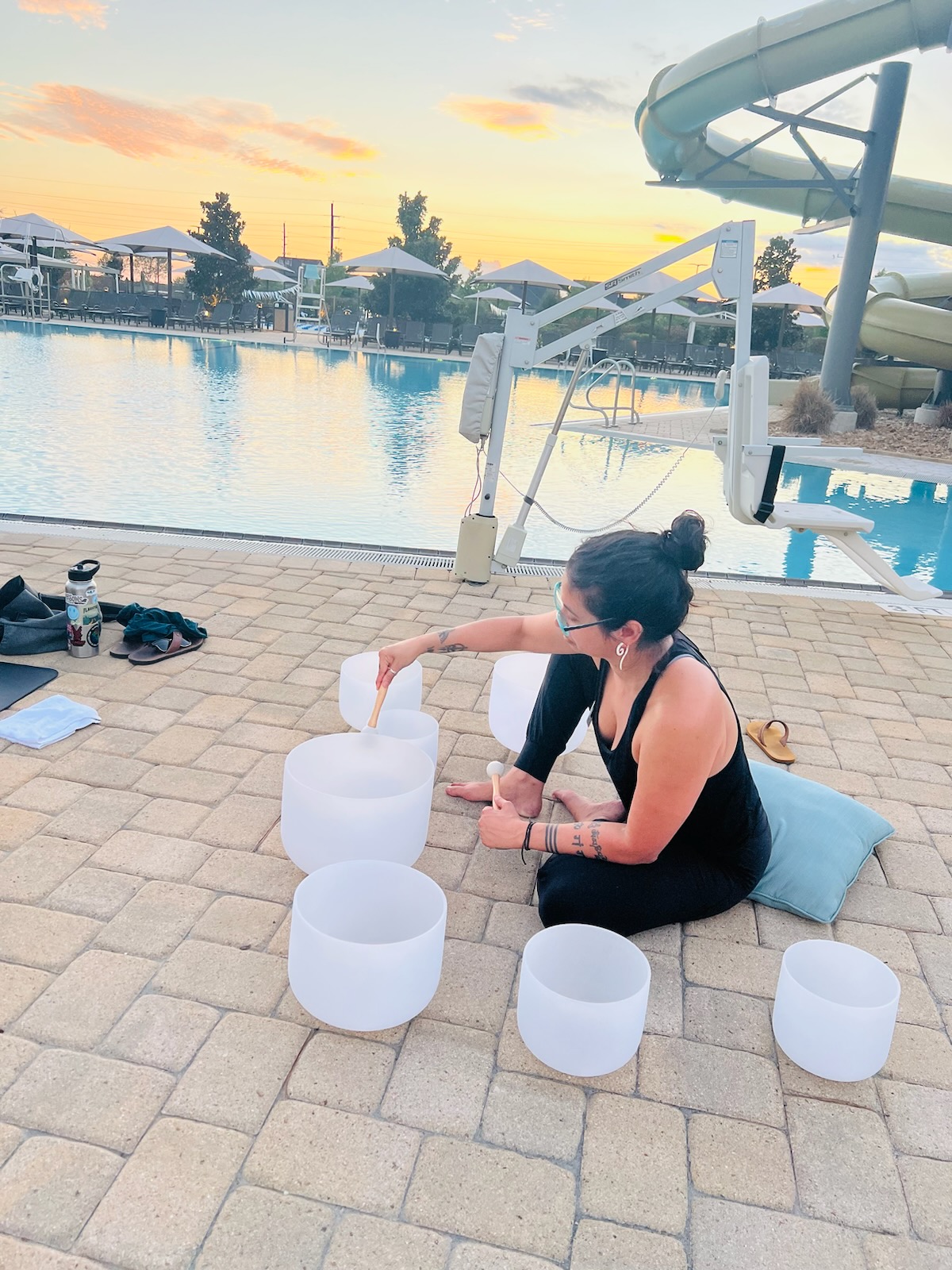 Woman sitting on a cushion by a pool, playing singing bowls, with water slide and pool in background.