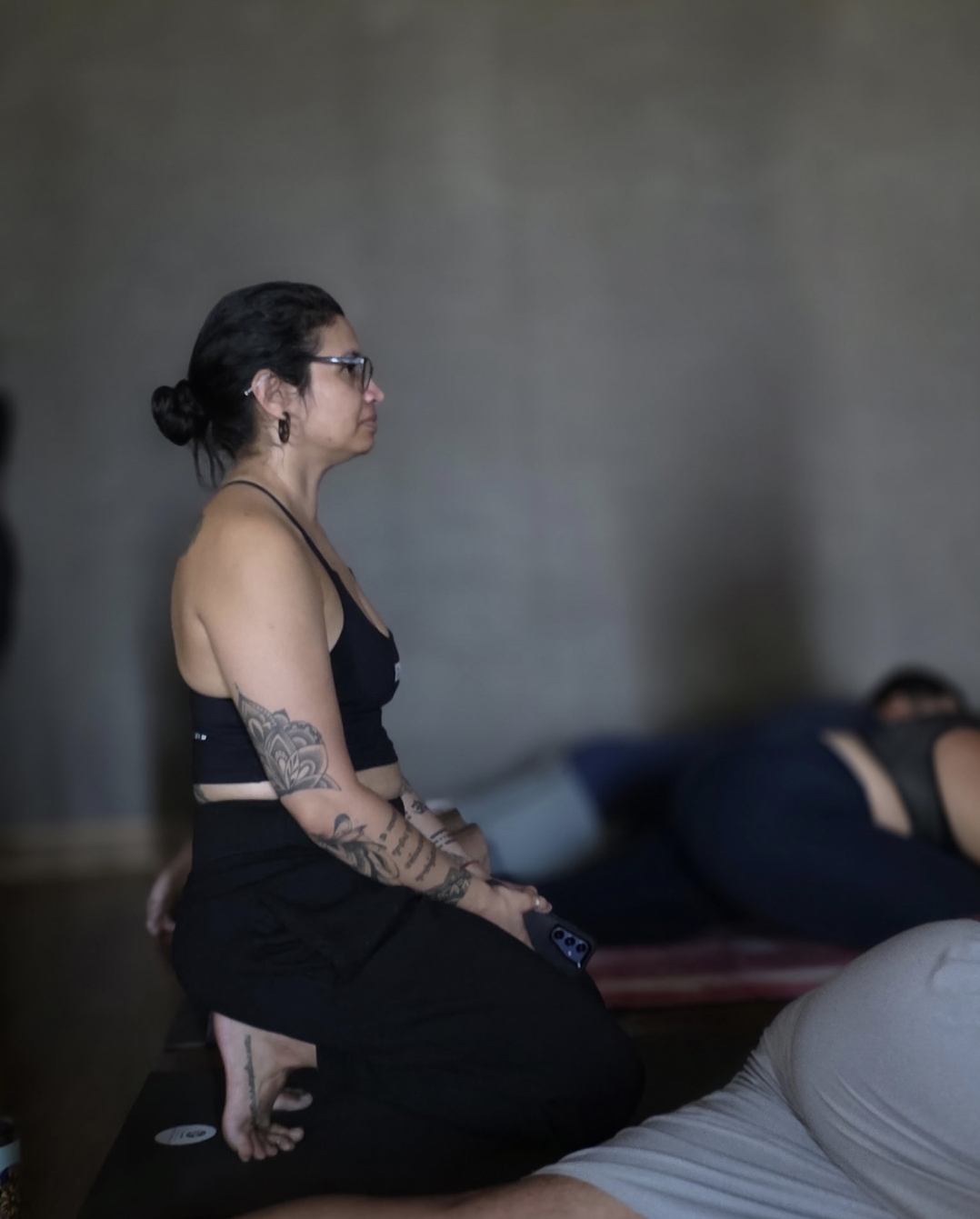 Woman with glasses and tattoos sitting cross-legged, with a person lying down in the background.