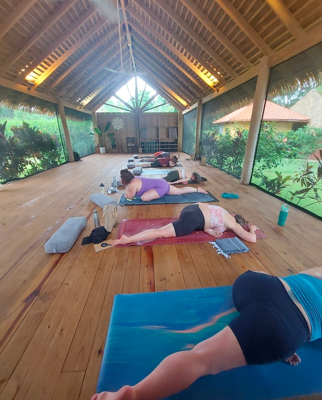 Group of people practicing yoga on mats in a wooden, open-air pavilion with scenic wall murals, natural light, and a high ceiling.