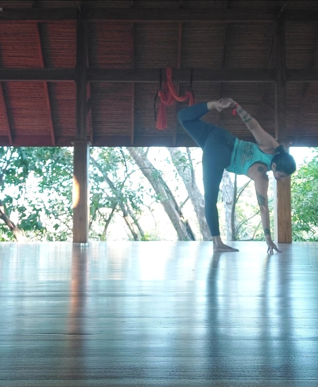 Person performing a handstand with one leg bent, under a wooden roof with trees outside, on a polished floor.