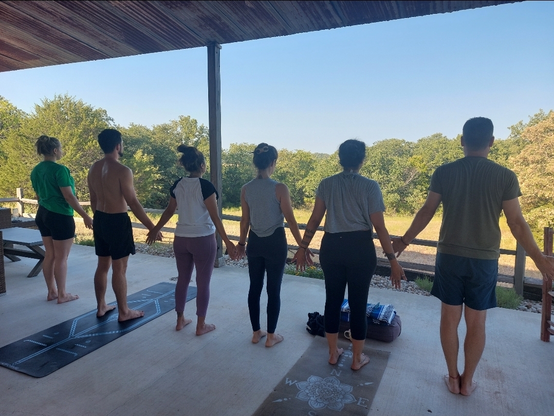 Seven people stand on a porch holding hands, facing a scenic outdoor view with trees and blue sky.