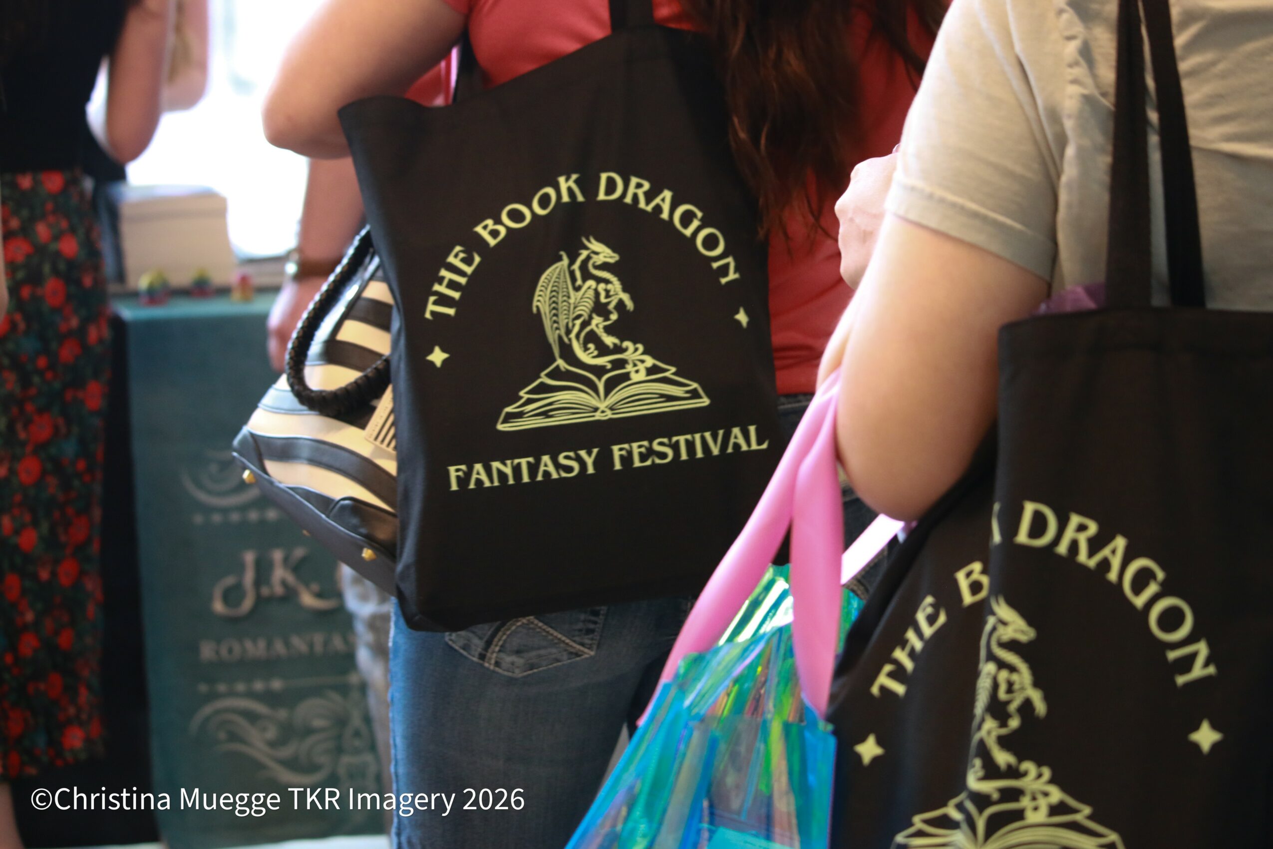 People holding black tote bags with yellow text and a dragon logo at a fantasy festival, some with pink and blue umbrellas.