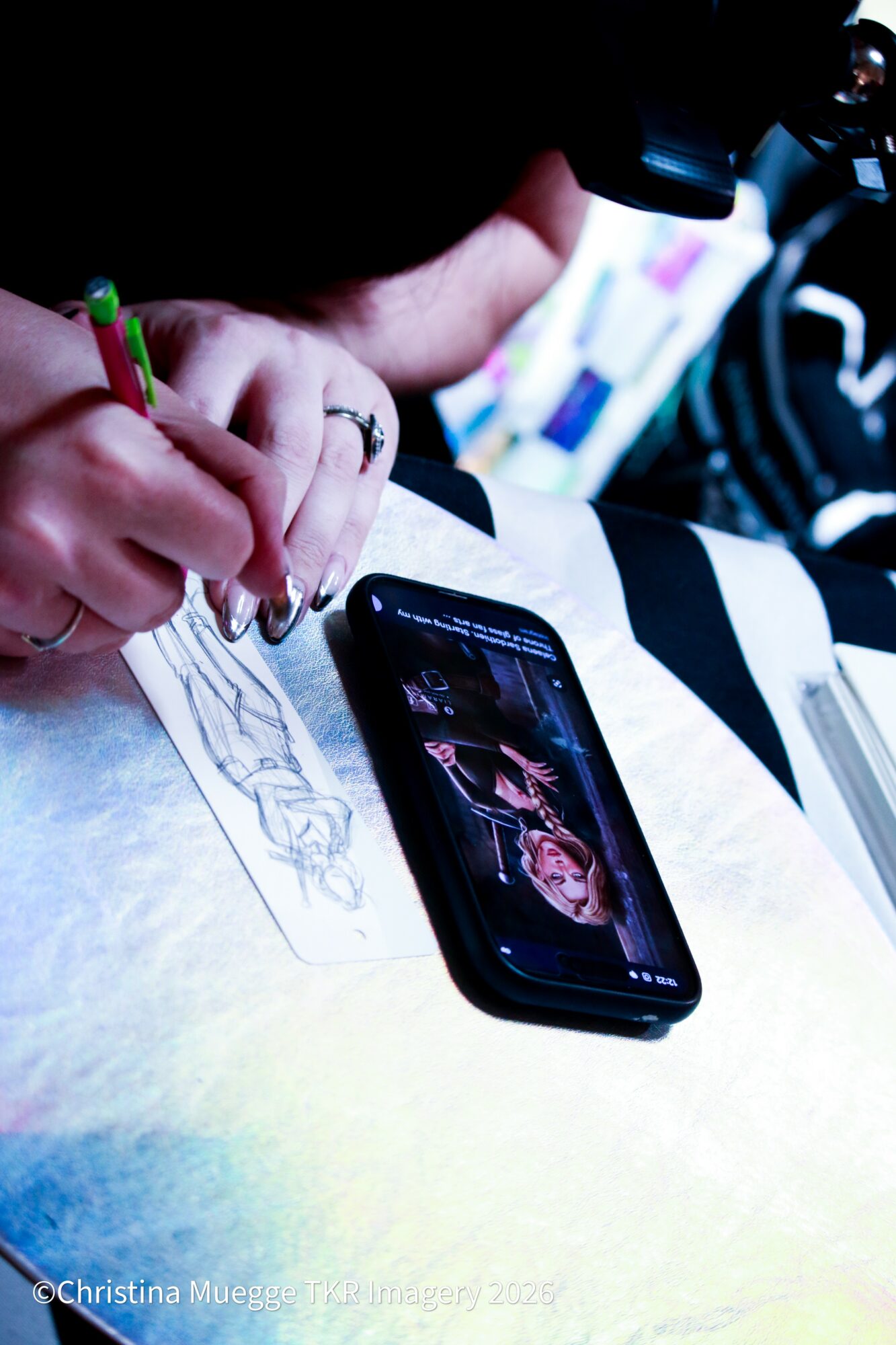 Person drawing on paper with a smartphone nearby, in a well-lit environment. Hand holding a green pen, focus on drawing process.