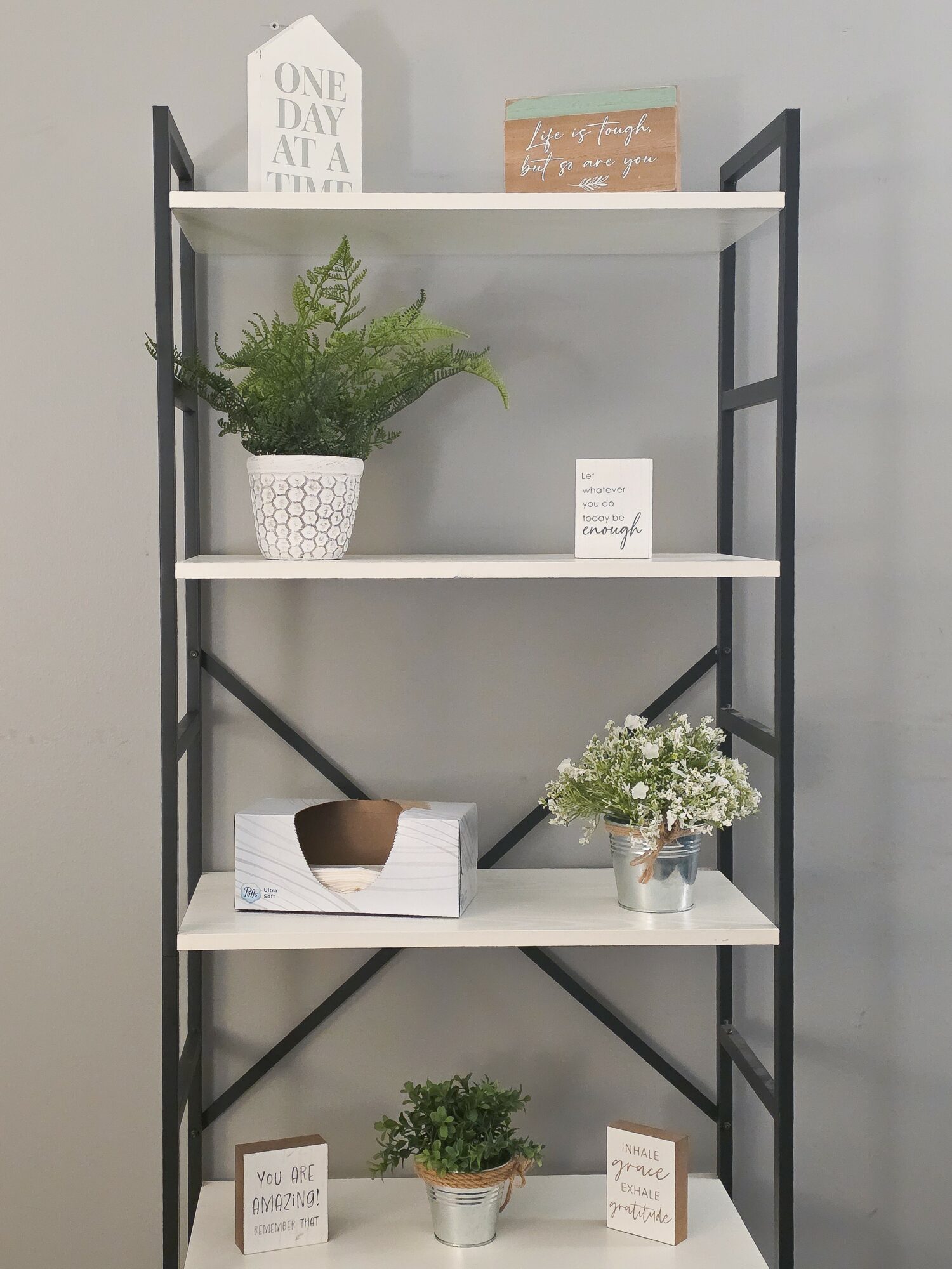 Black metal shelf with four white shelves holding plants, signs, and decorative items against a gray wall.