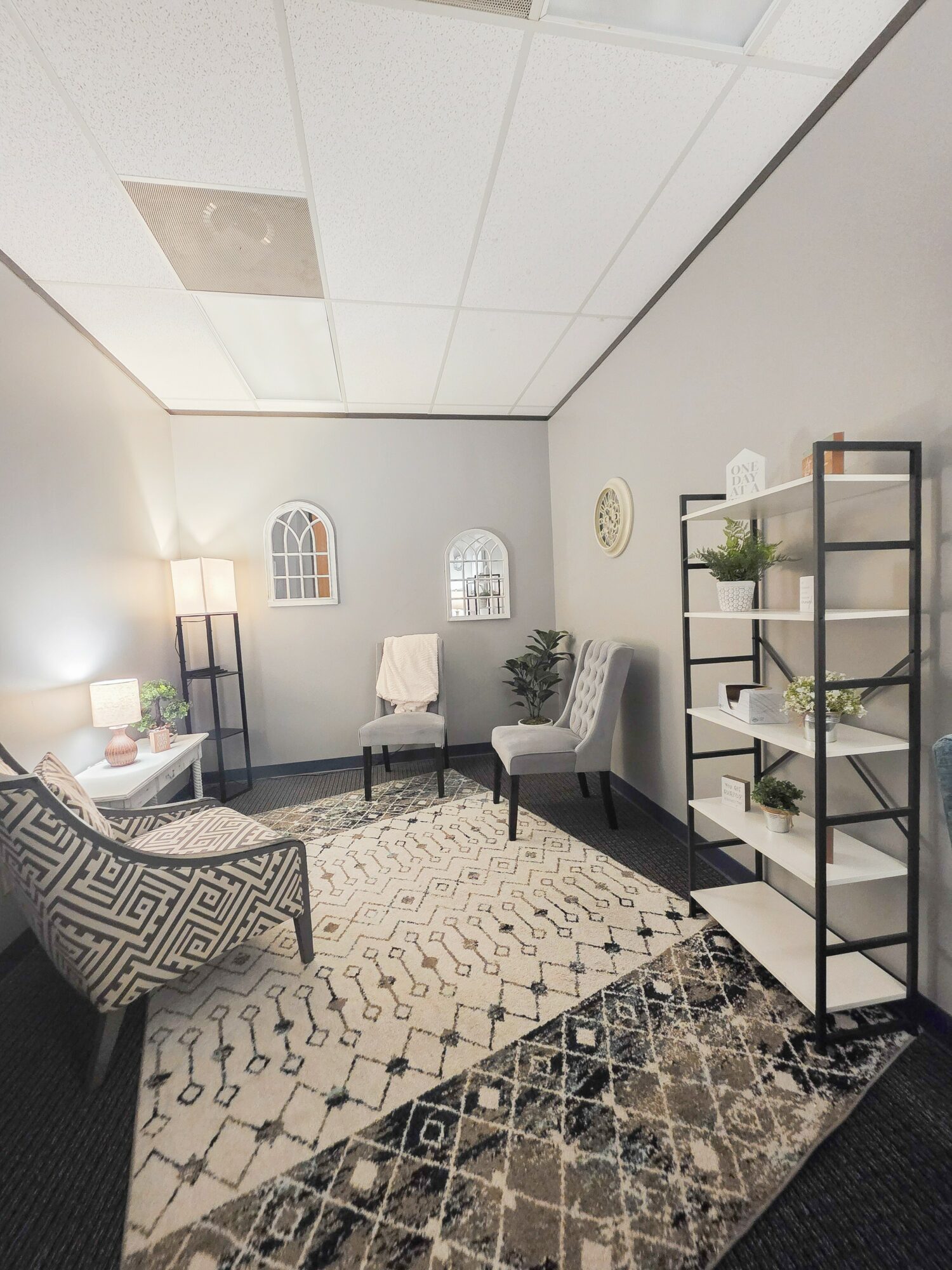 Living room with chairs, a patterned armchair, a black metal shelf with plants, and a rug, ceiling with square tiles.