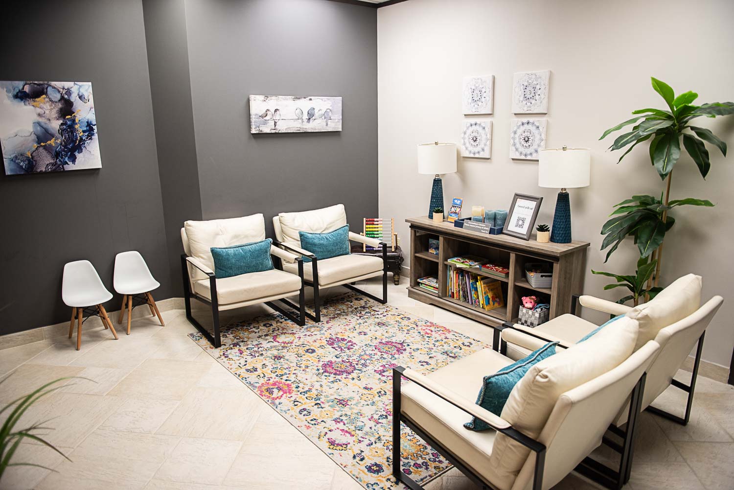 Living room with white and dark gray walls, two white armchairs, two white chairs, a bookshelf, and artwork, with a colorful rug.
