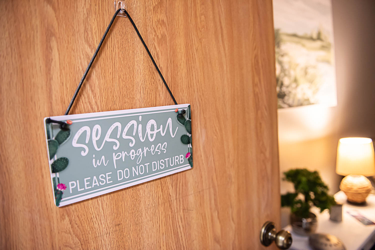 A sign hanging on a wooden wall reads 'session in progress, please do not disturb' with decorative leaves and flowers, and a blurred background with a lamp and plant.