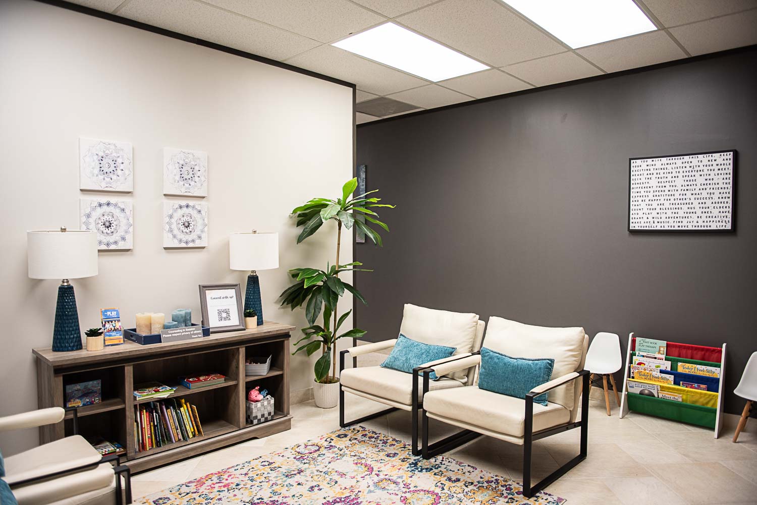 Office room with white and dark gray walls, two white chairs with blue cushions, a bookshelf, and framed pictures.