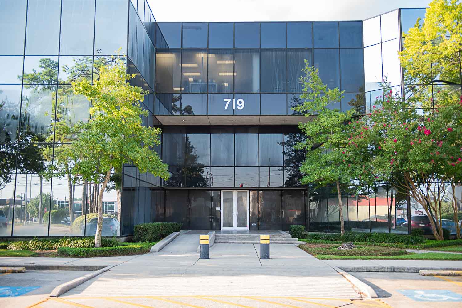 Modern glass building with the number 719 above the entrance, surrounded by trees and a sidewalk.