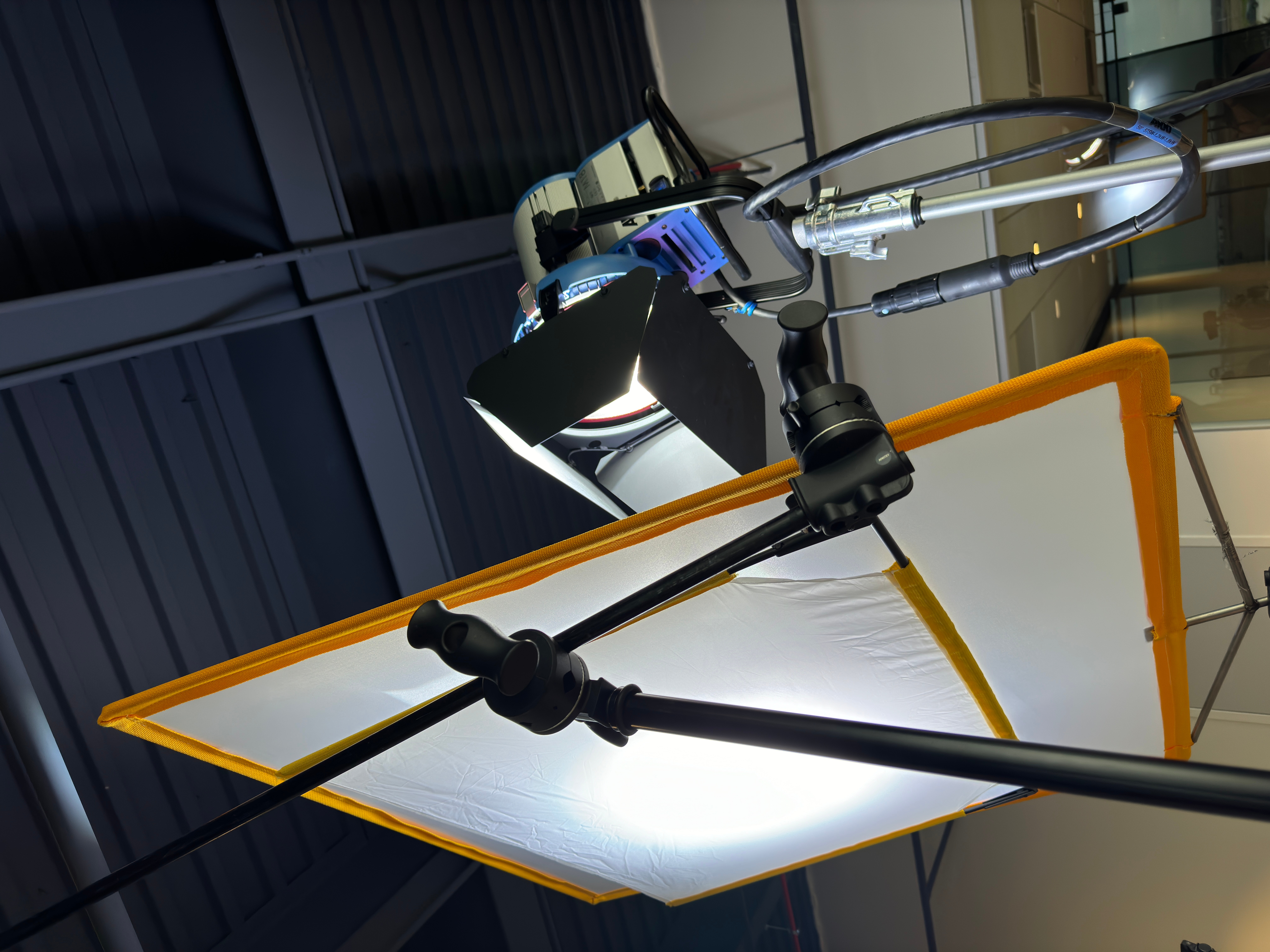 Lighting equipment and a reflector are set up indoors, with a dark ceiling and glass windows in the background.