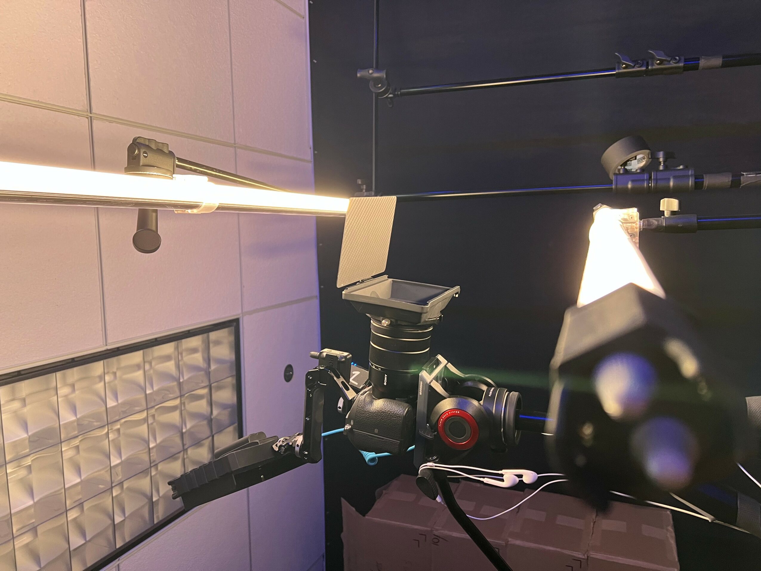 Camera setup with a black mount and a lens, positioned in a room with purple and black walls, ceiling lights, and a glass block window.