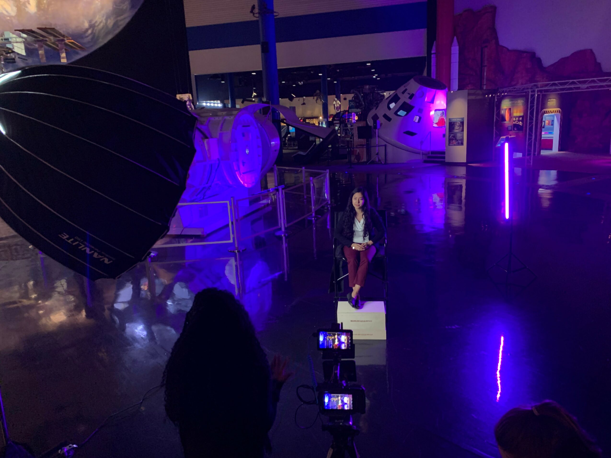 Person taking photo of a display with a woman standing on a platform, illuminated by purple lights, in an indoor space.