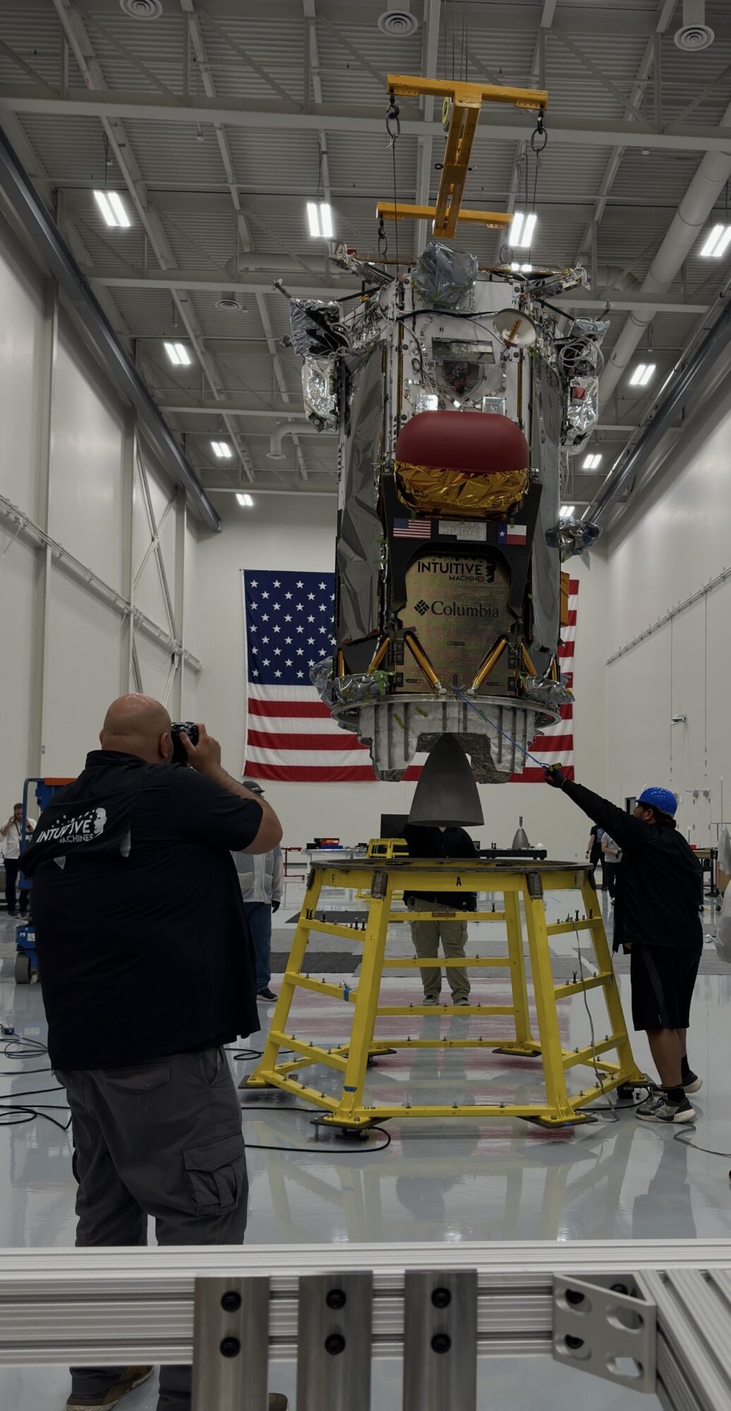 People working on a large spacecraft inside a hangar with an American flag in the background.