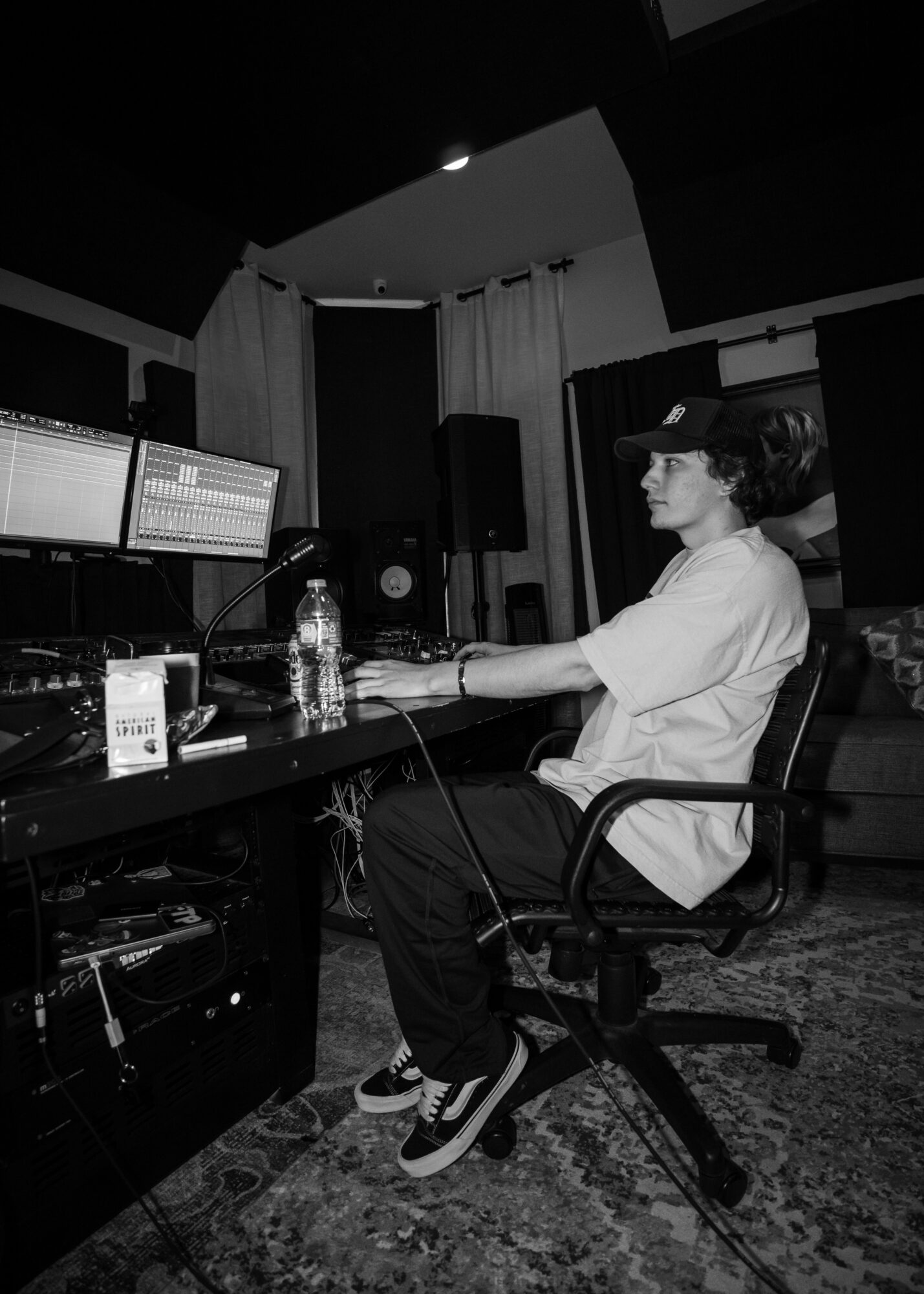 Person sitting at a desk in a recording studio with audio equipment and computer screens, wearing headphones and a white shirt.