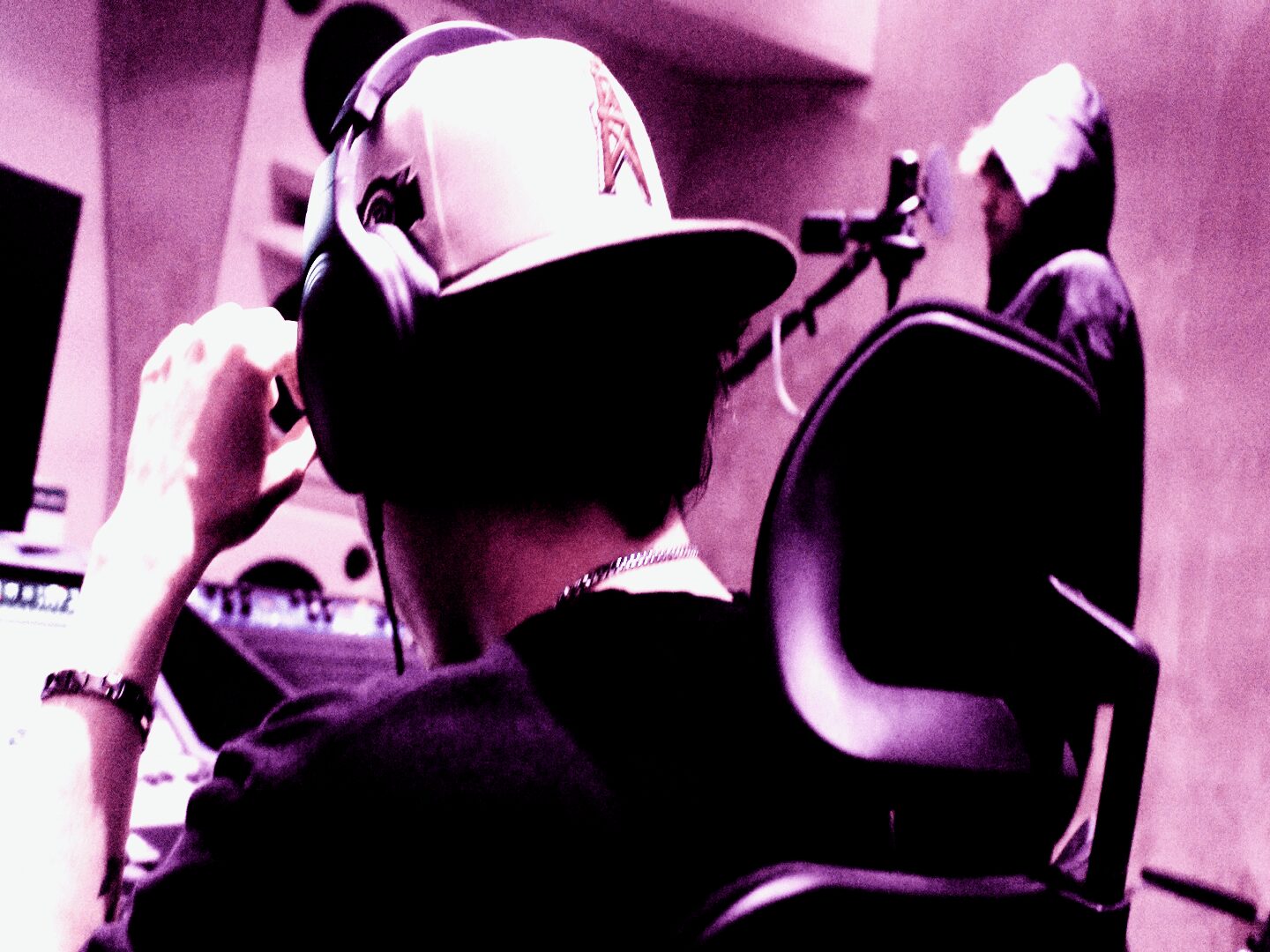 Person wearing headphones and a cap in a recording studio, with another person in the background.