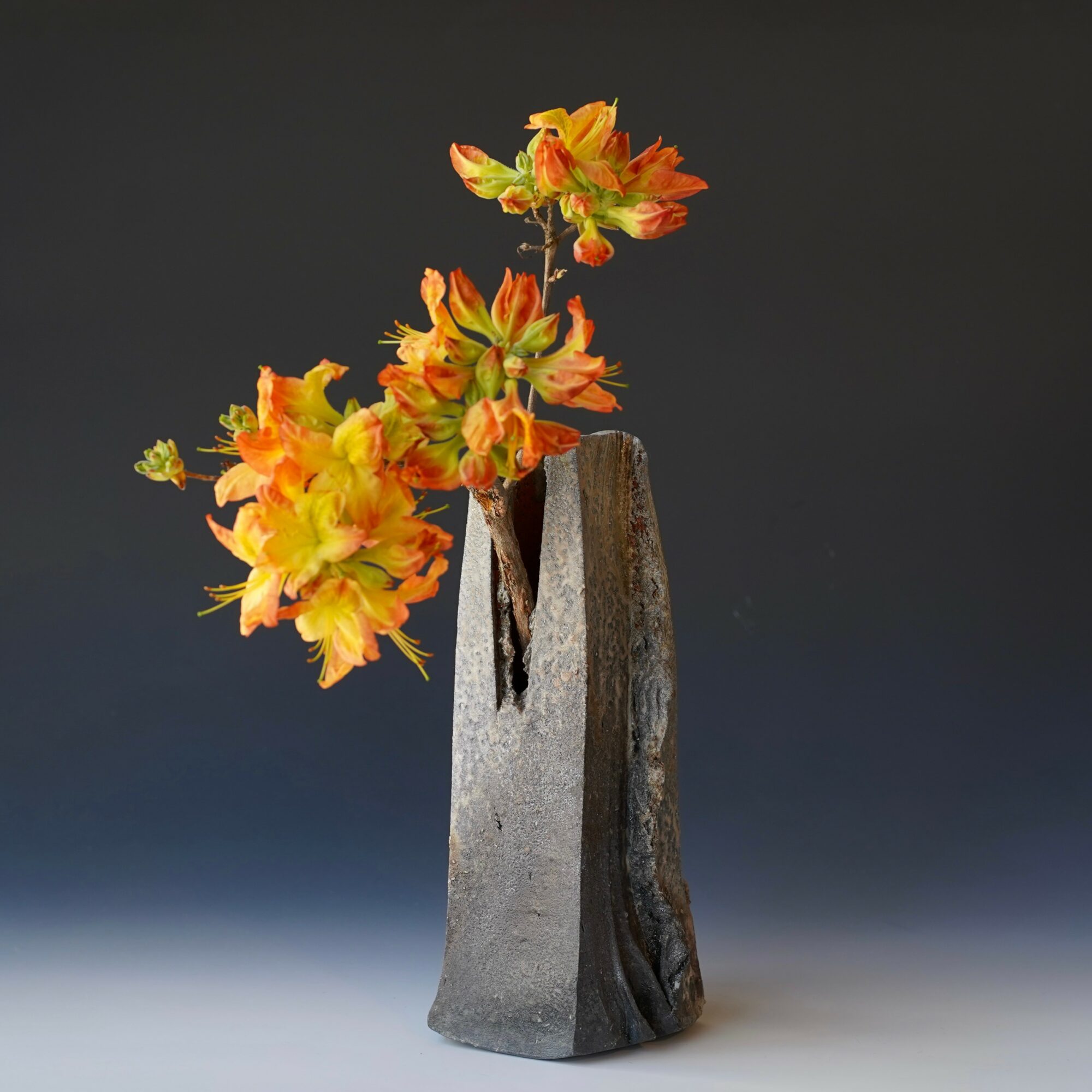 A tall, textured gray vase with orange and yellow flowers against a dark background.