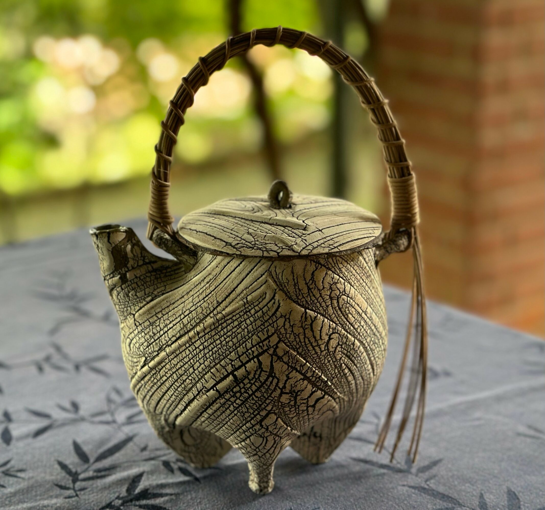 Decorative teapot with a textured, wood-like surface and a woven handle, placed on a tablecloth with a blurred outdoor background.