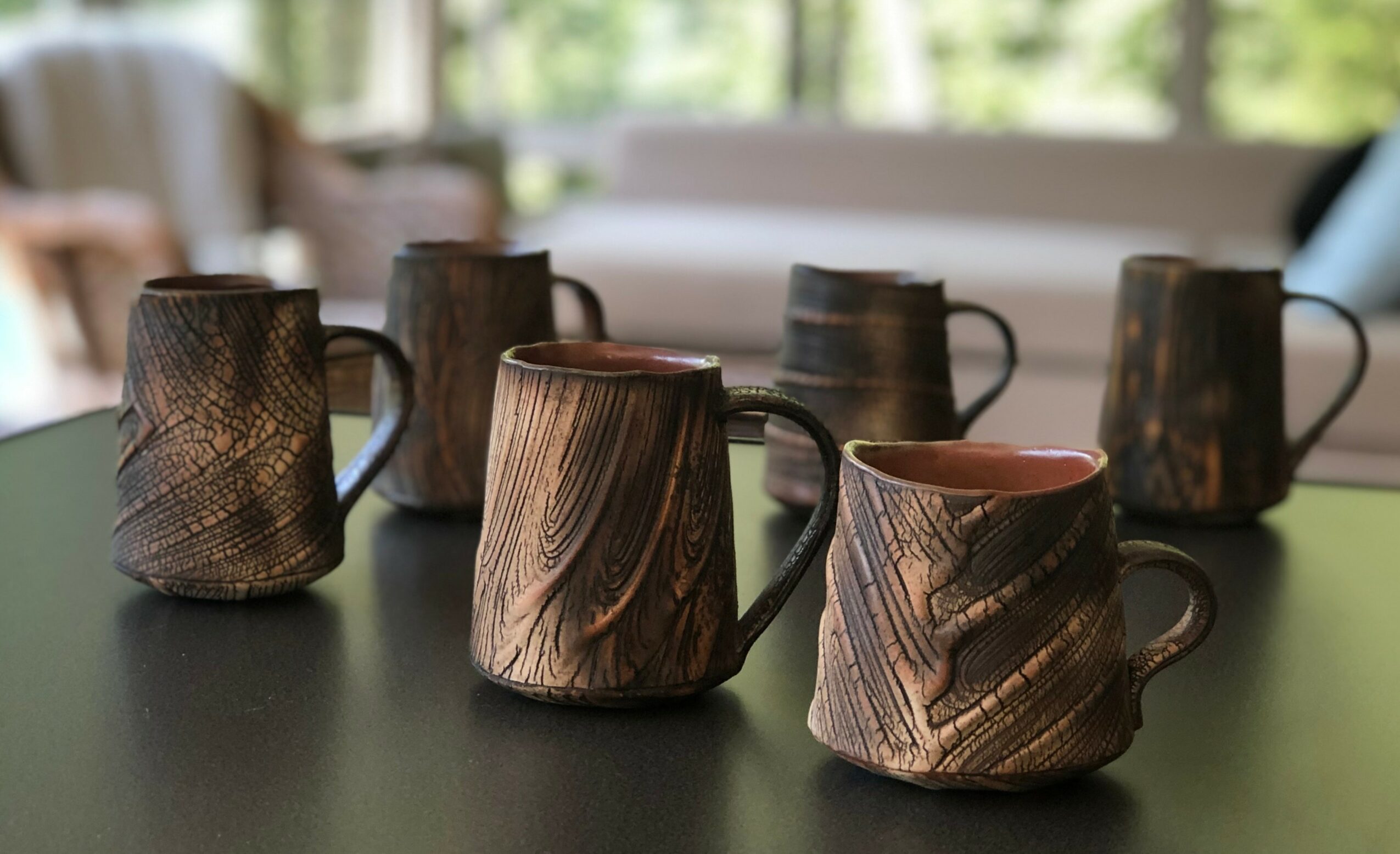 Six ceramic mugs with wood grain patterns on a dark table, background of a bright room with windows and furniture.