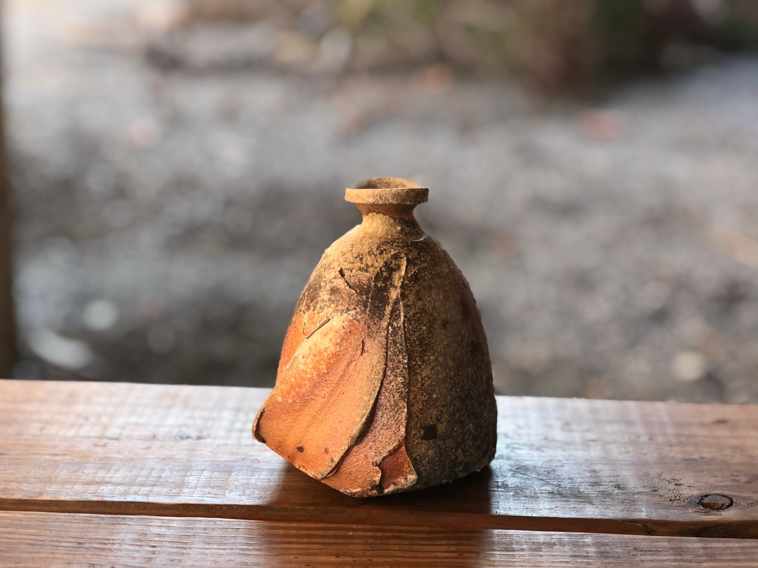 Small, weathered clay pot with a narrow opening, placed on a wooden surface outdoors.