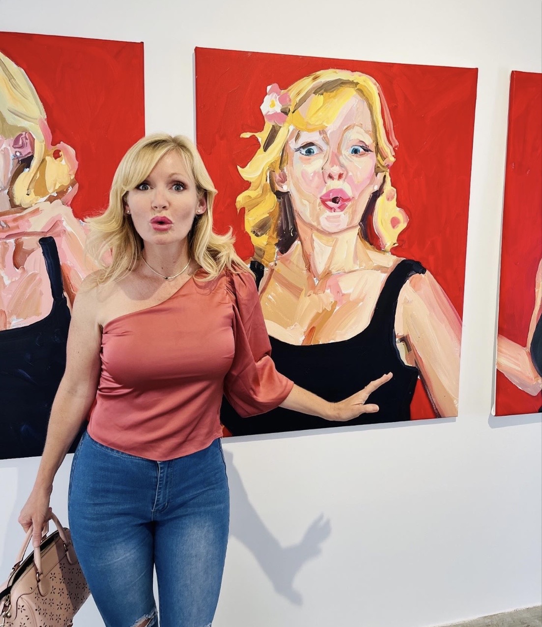 Woman with blonde hair in a pink top and blue jeans standing in front of colorful portrait paintings, pointing at one painting.