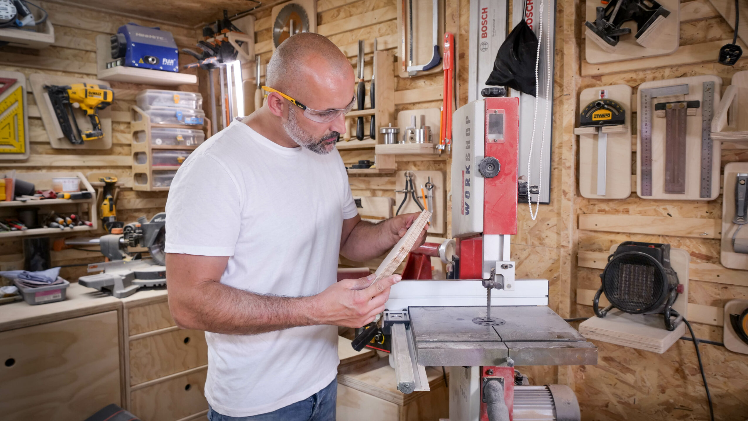 Man in workshop examining a piece of wood, surrounded by tools and wooden walls.