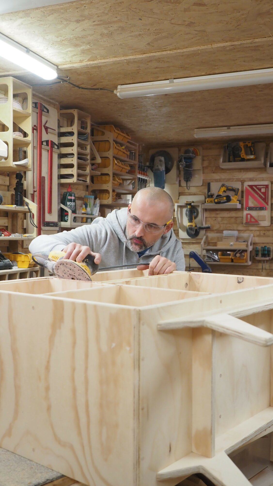 Person working at a woodworking shop with tools and wooden structures, viewed from the side, with shelves and equipment in the background.