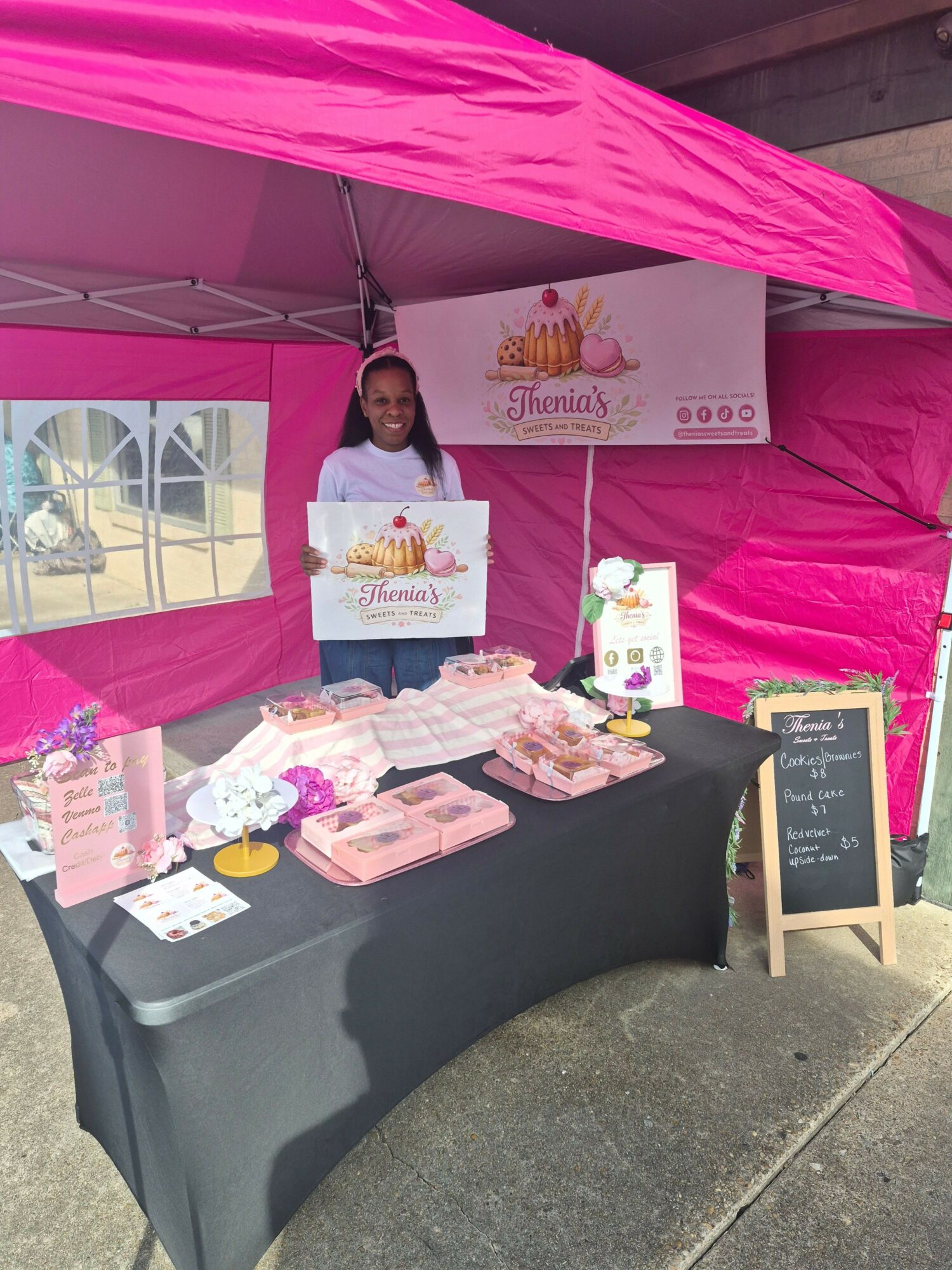 Person standing behind a table with pink decorations, promotional materials, and a sign, under a pink canopy.